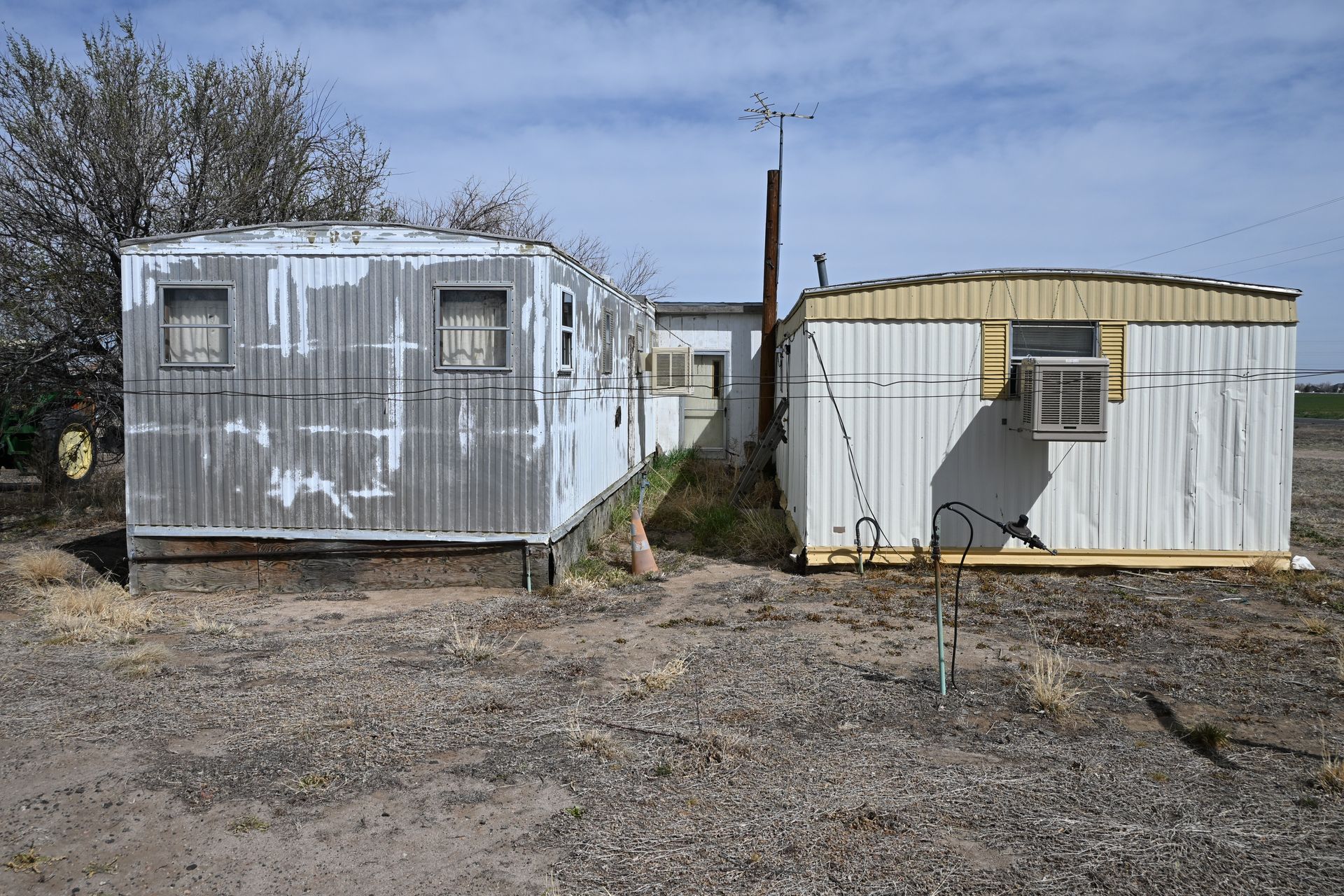 Two small weathered mobile homes in a gravel lot, one white and one beige, with sparse trees behind them.