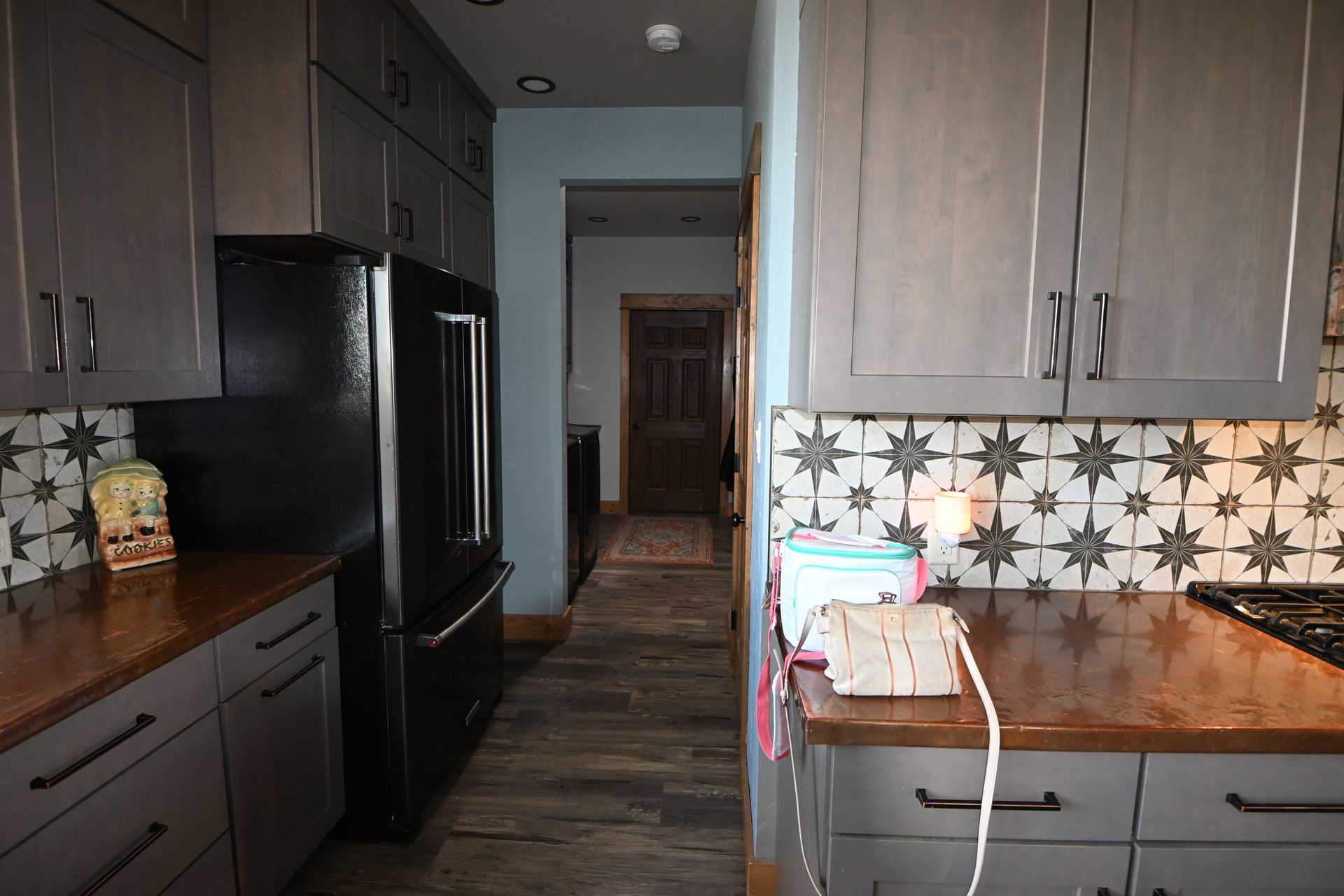 Narrow kitchen hallway with dark cabinets, black fridge, patterned backsplash, and wood countertops.