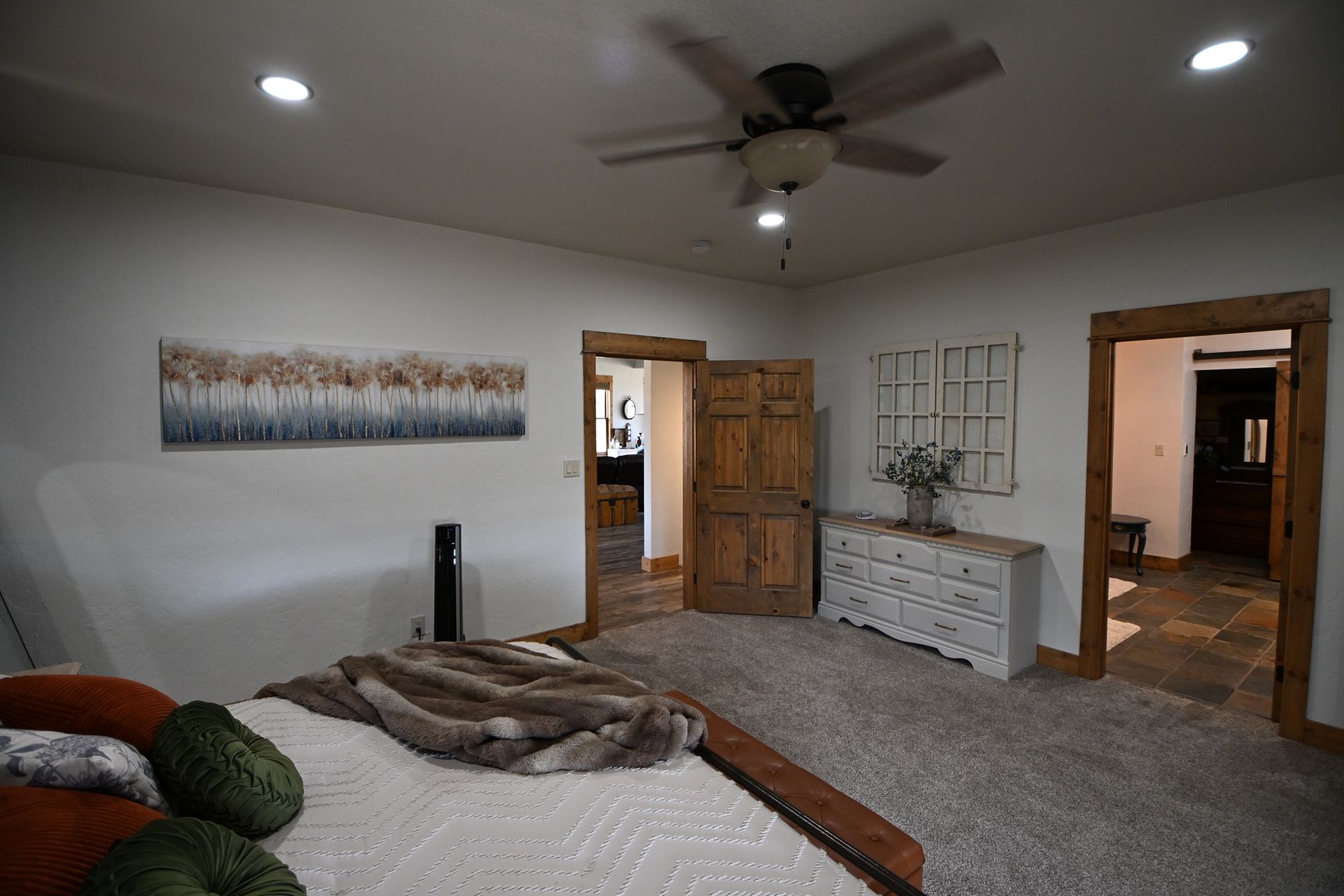 Spacious bedroom with a bed, ceiling fan, dresser, and open wooden doors leading to a hallway