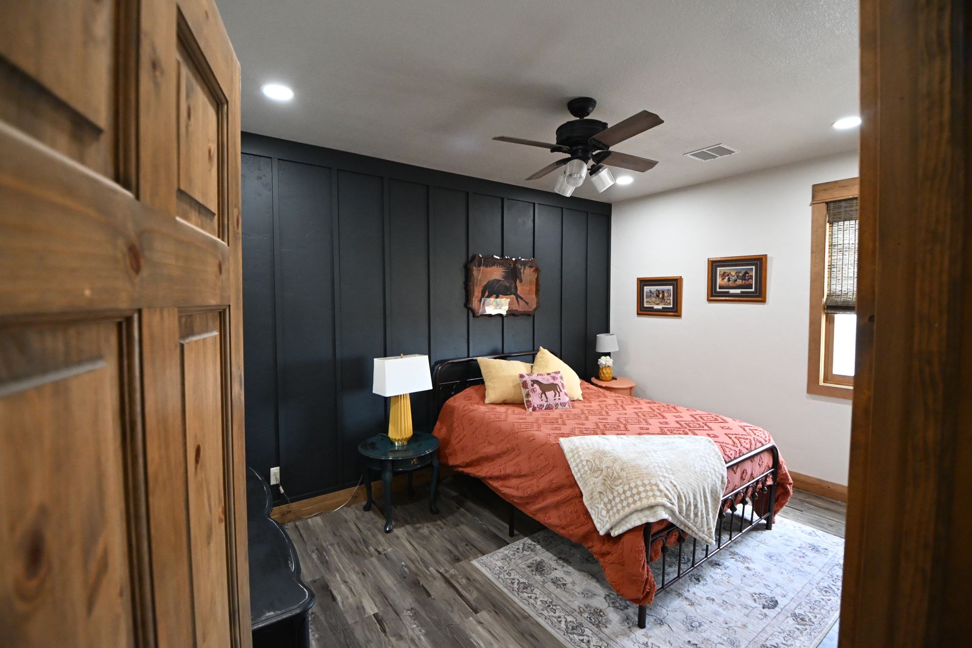 Modern bedroom with black accent wall, orange bedspread, gray rug, ceiling fan, and wooden doors.