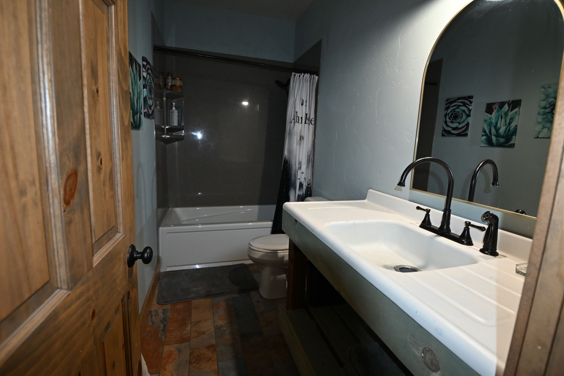 Dim bathroom with a freestanding tub, long white vanity sink, dark wood floor, and wooden door