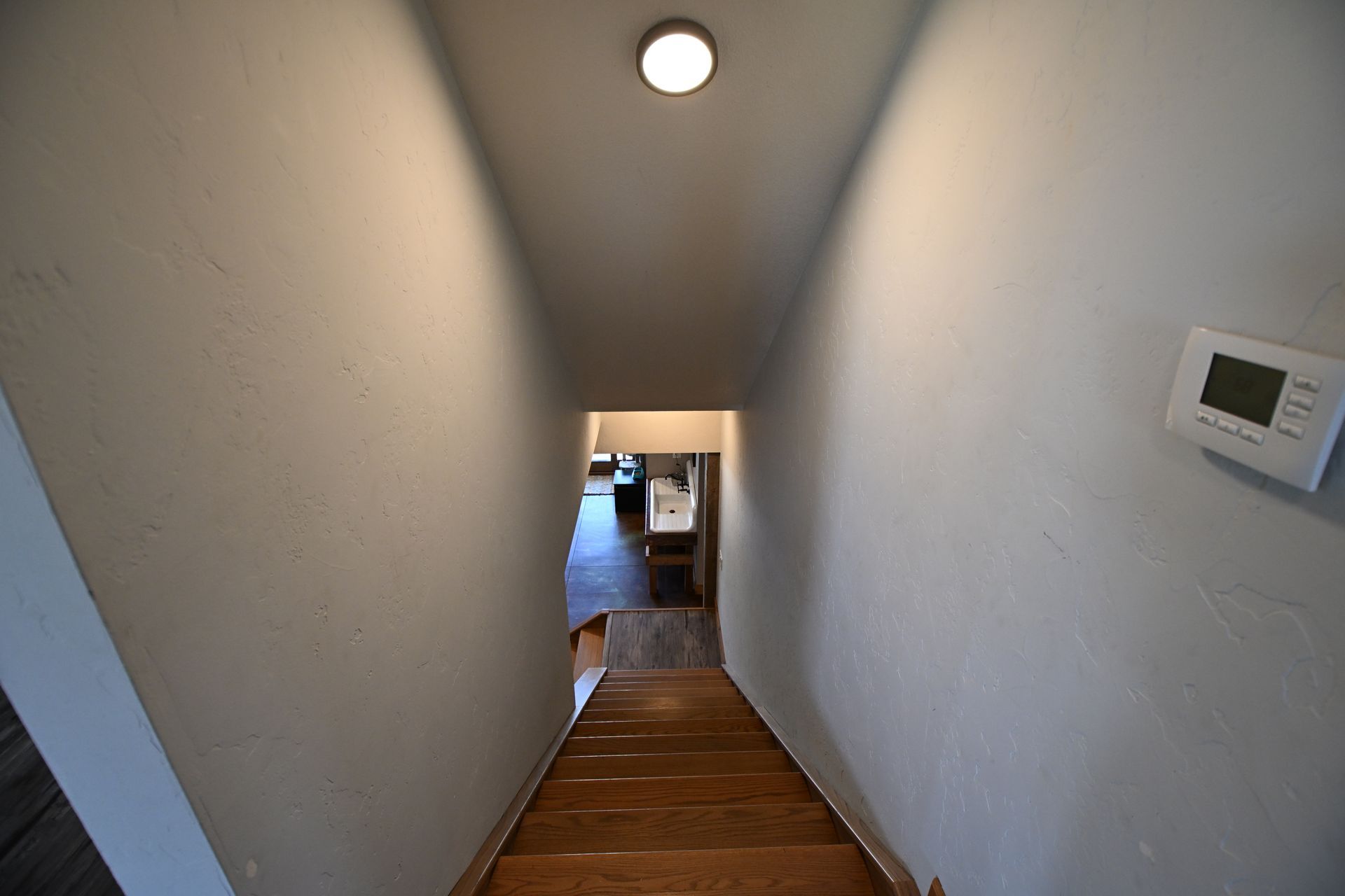 Narrow stairwell with white walls, wood steps, ceiling light, and thermostat on the right.