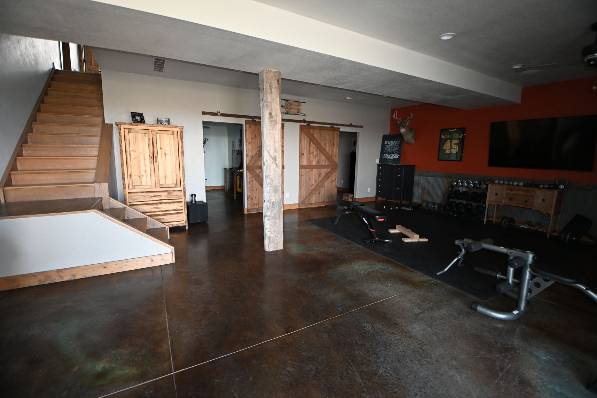 Empty basement gym with staircase, wooden doors, weight bench, and exposed support beam