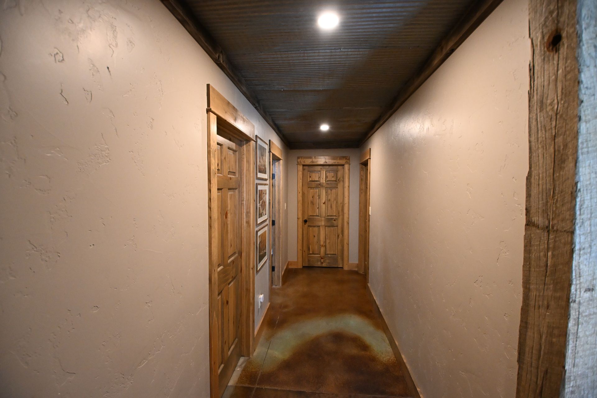 Narrow indoor hallway with beige walls, wood doors, dark ceiling, and a light at the far end