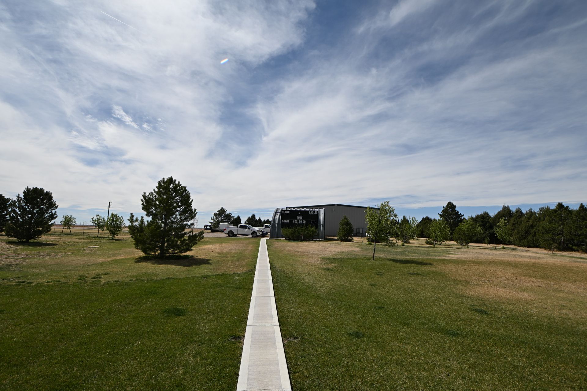 Wide grassy field with a white path leading to a building under a cloudy blue sky.