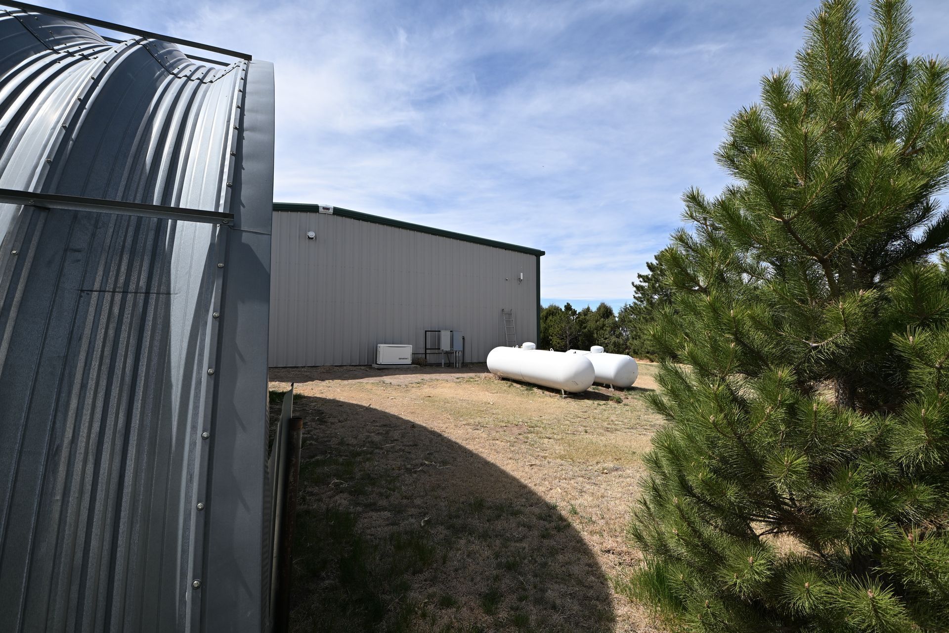 Industrial building with silver tank, white propane tanks, and a tree beside a dirt lot under a blue sky