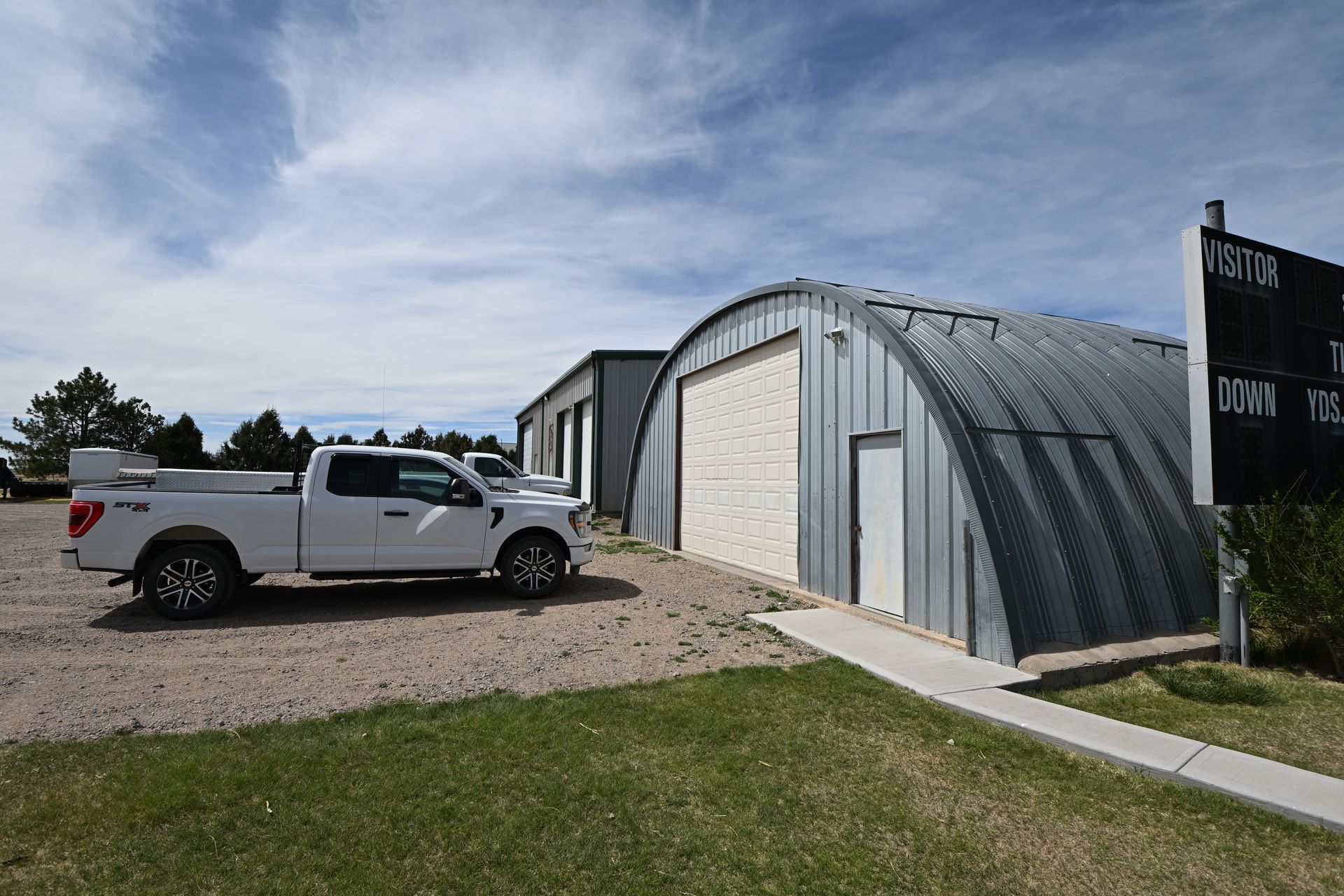 White pickup truck parked beside a curved metal building on a gravel lot under a blue sky