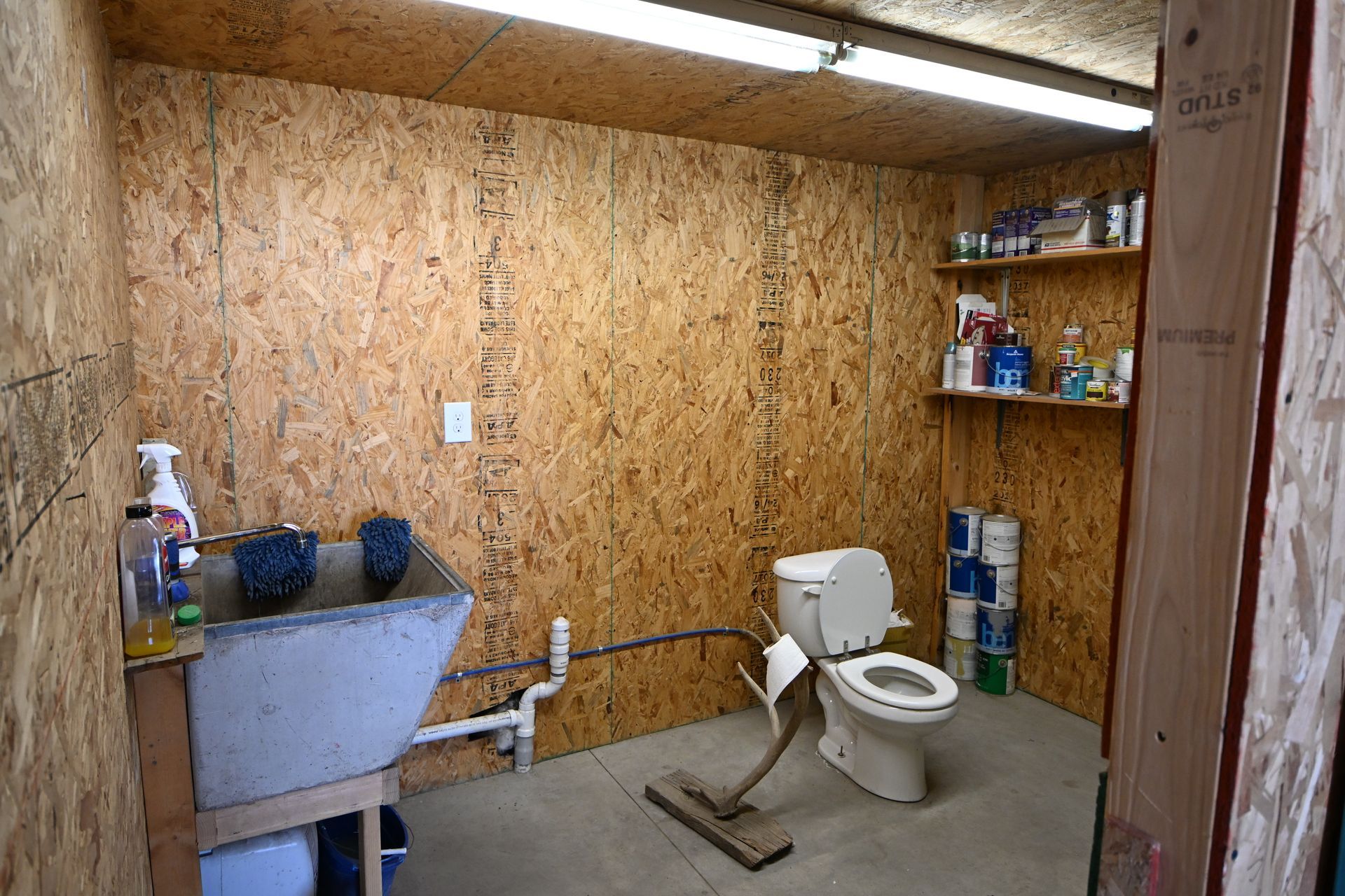 Small unfinished bathroom with toilet, sink, exposed plywood walls, and a utility shelf.