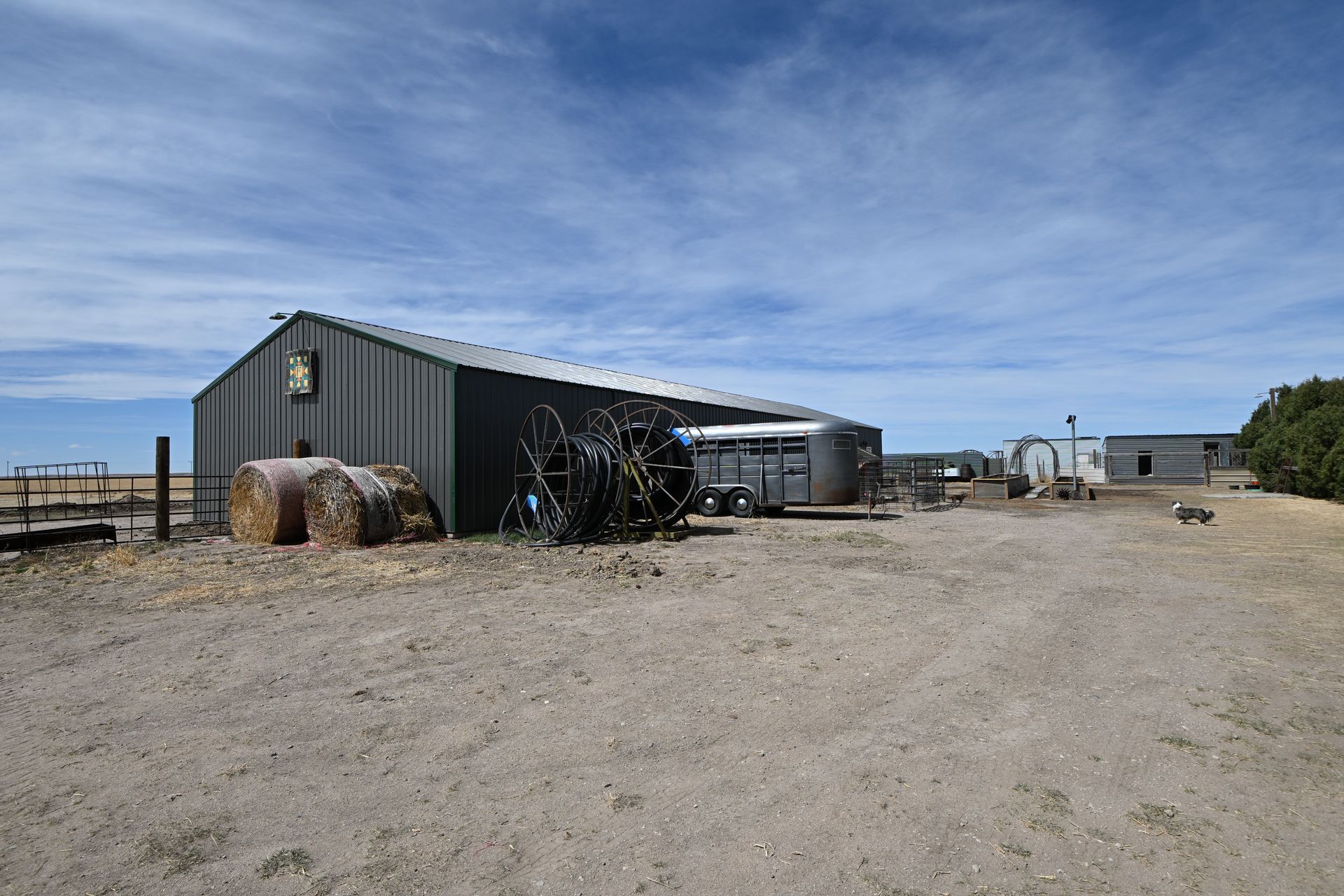 Rural metal shed in a dusty yard under a blue sky, with hay bales and farm equipment nearby
