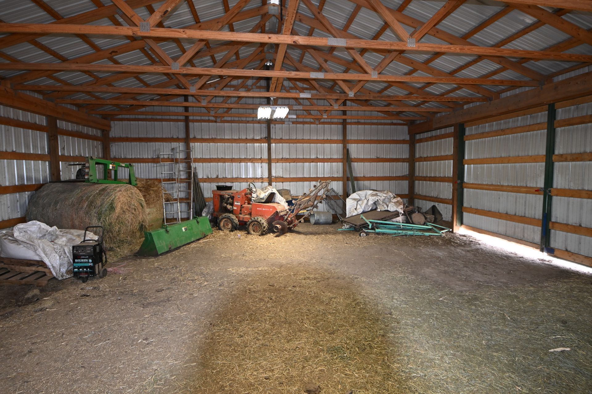 Inside a barn, farm equipment and supplies are stacked along the walls under a wooden truss roof.