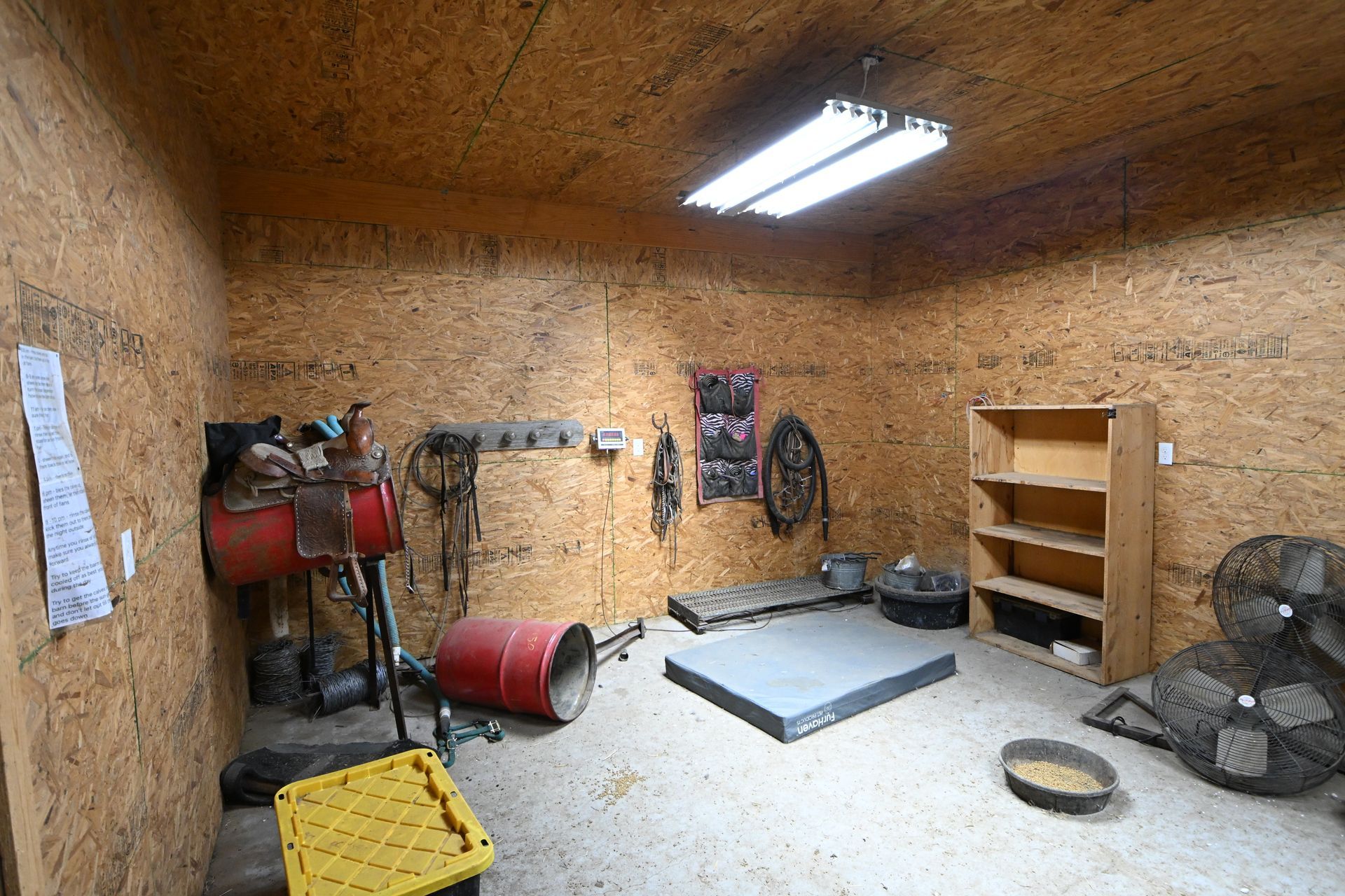 Workshop with wood-paneled walls, fluorescent lights, fan, shelves, tools, and equipment on the floor.