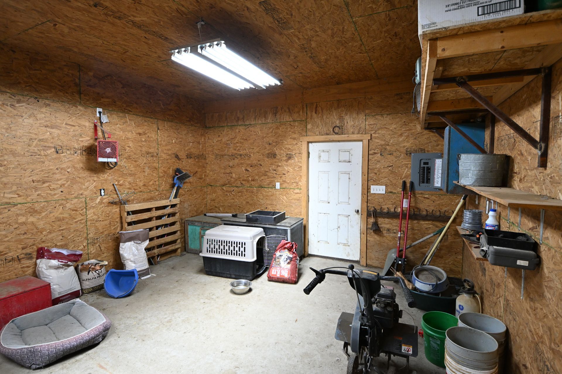 Wood-paneled garage with a white door, overhead lights, tools, and storage items scattered around.