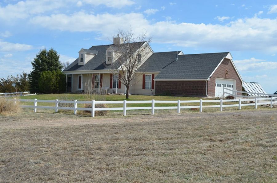House with white fence, garage, and dormers on a sunny day in a field.