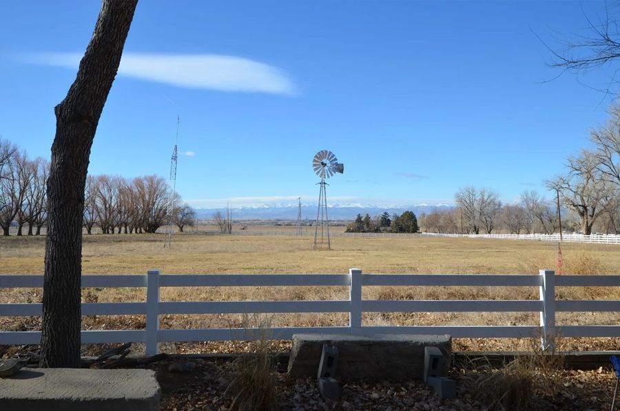 Field with white fence, windmill, bare trees, and a bright blue sky.