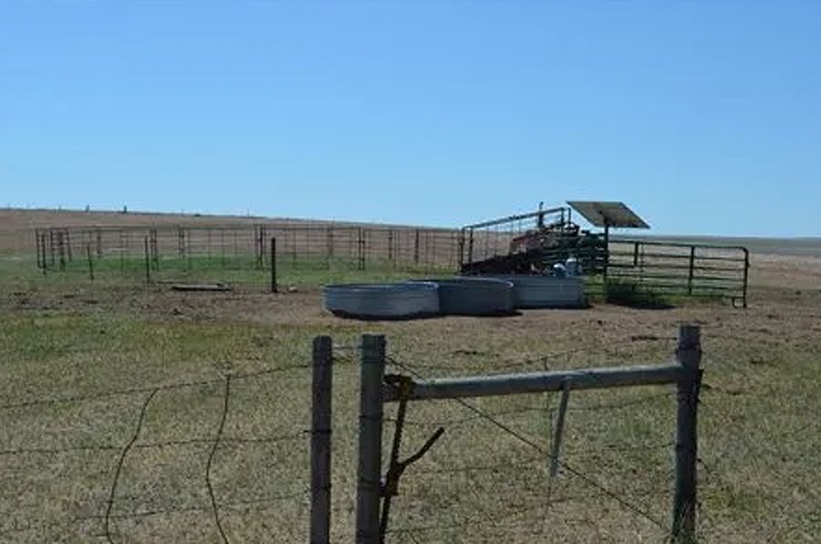 A rural scene with a weathered fence, metal water troughs, and a tractor under a small shelter on a sunny day.