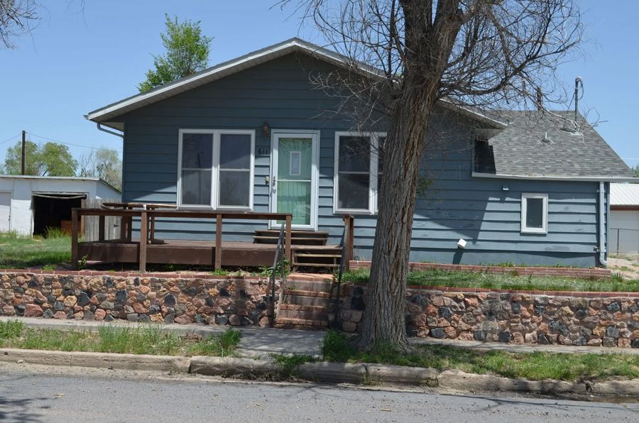 Blue house with a deck and stone retaining wall, trees, and a garage on a sunny day.