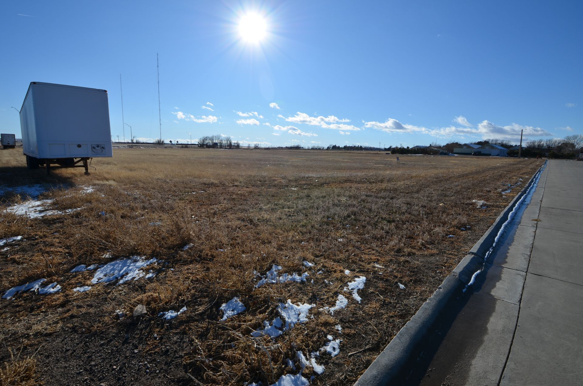 A sunny, open field with sparse vegetation, a white trailer, and a paved road.