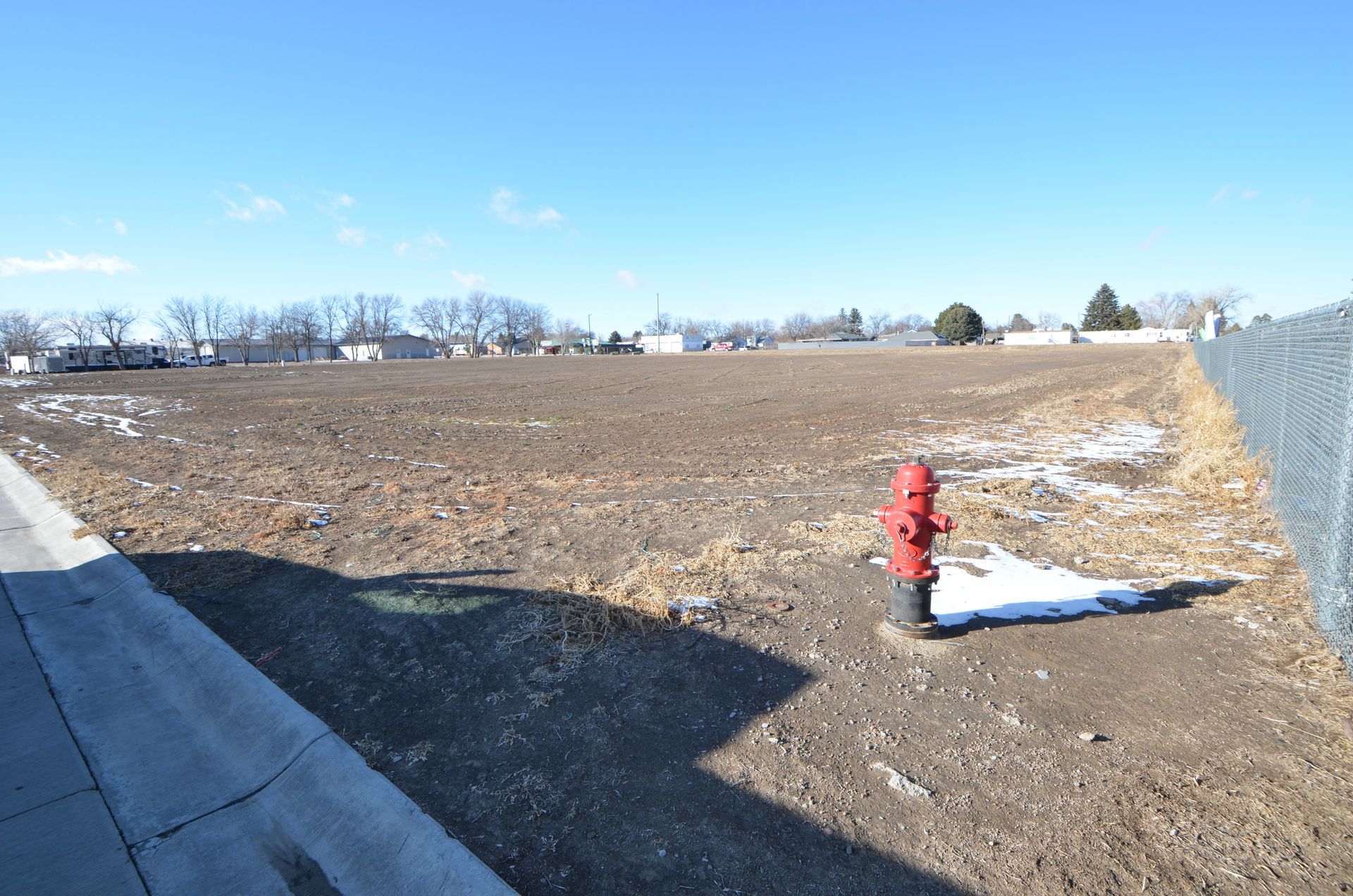 An empty dirt lot with a red fire hydrant, a sidewalk, and a chain-link fence on a sunny day.
