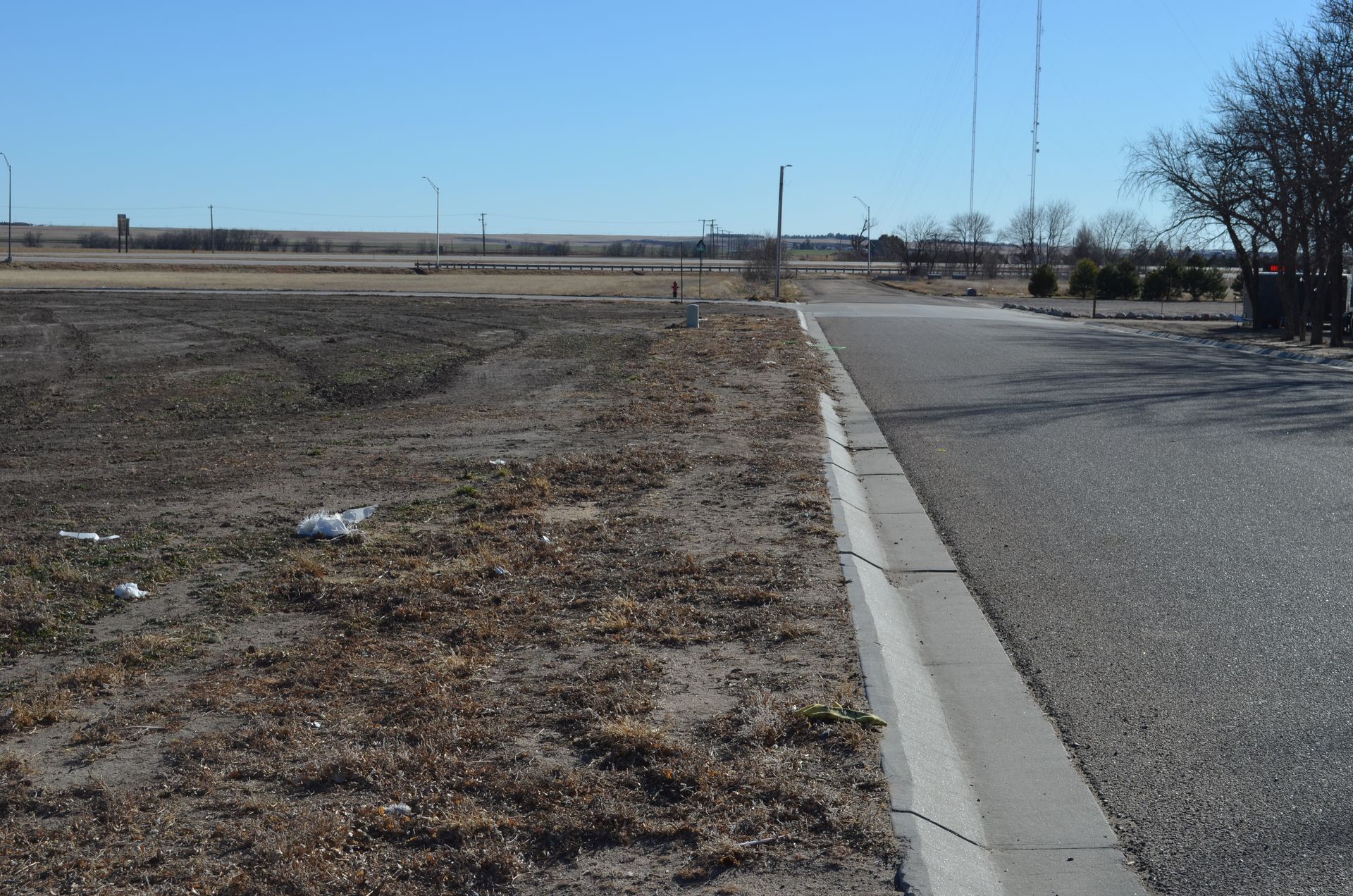 A paved road with a concrete curb borders dry grass and dirt. In the background are fields and a clear blue sky.