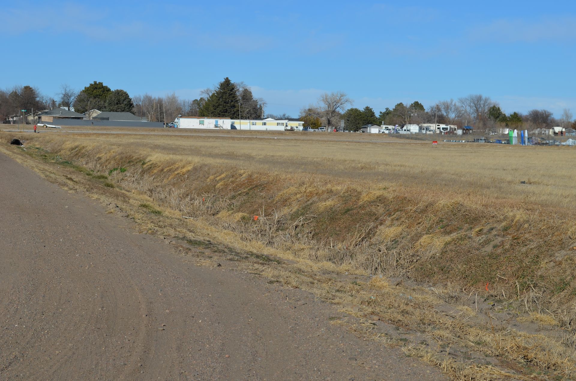 An empty, brown field with a dirt road in the foreground and houses in the distance under a blue sky.