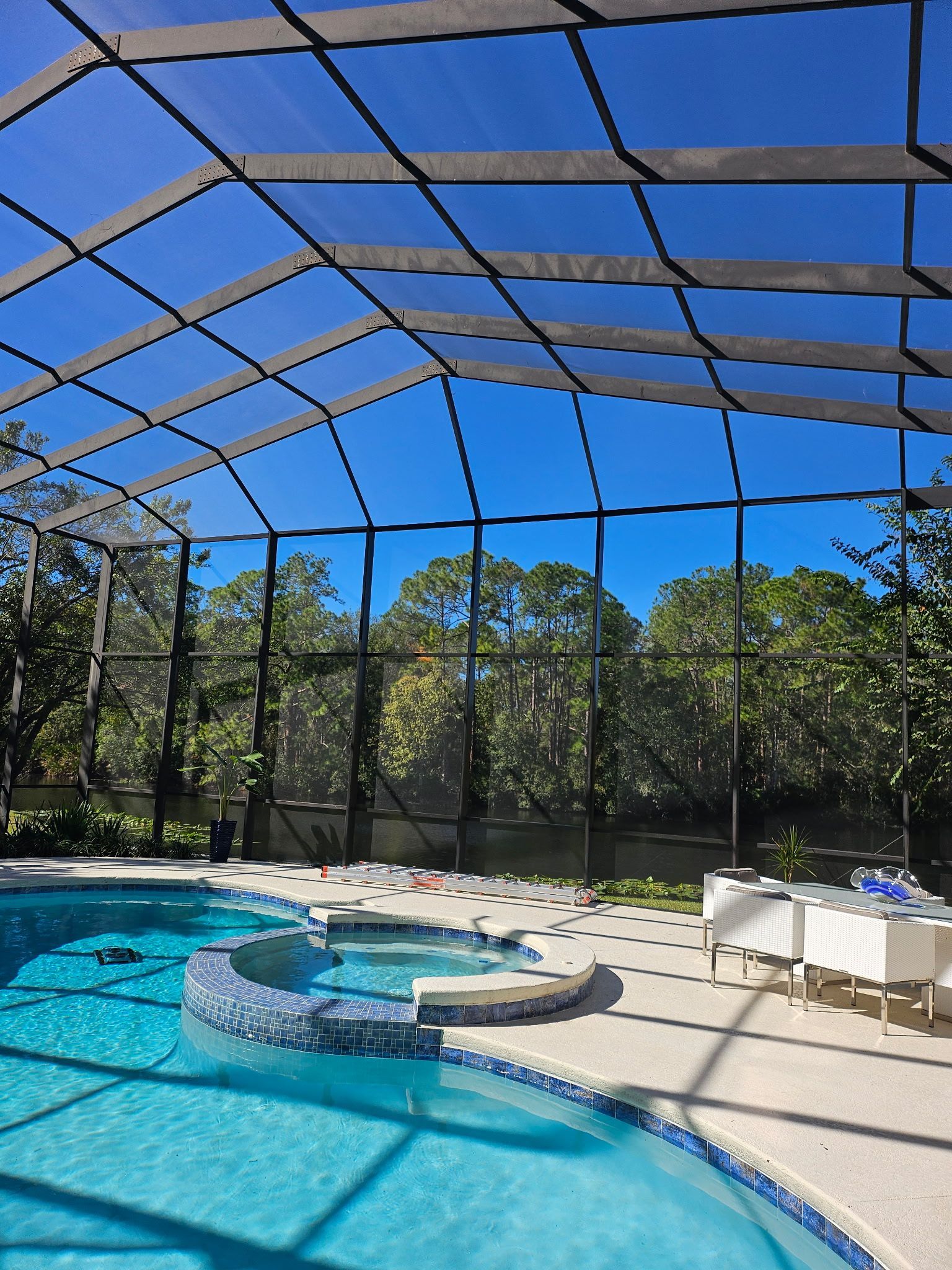 Screened-in backyard pool with a hot tub, white patio chairs, and a blue sky overhead