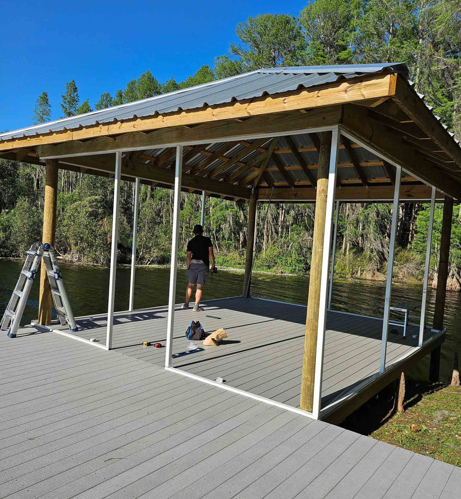 Partially built wooden pavilion on a gray deck, with tools and a person inside under a blue sky.