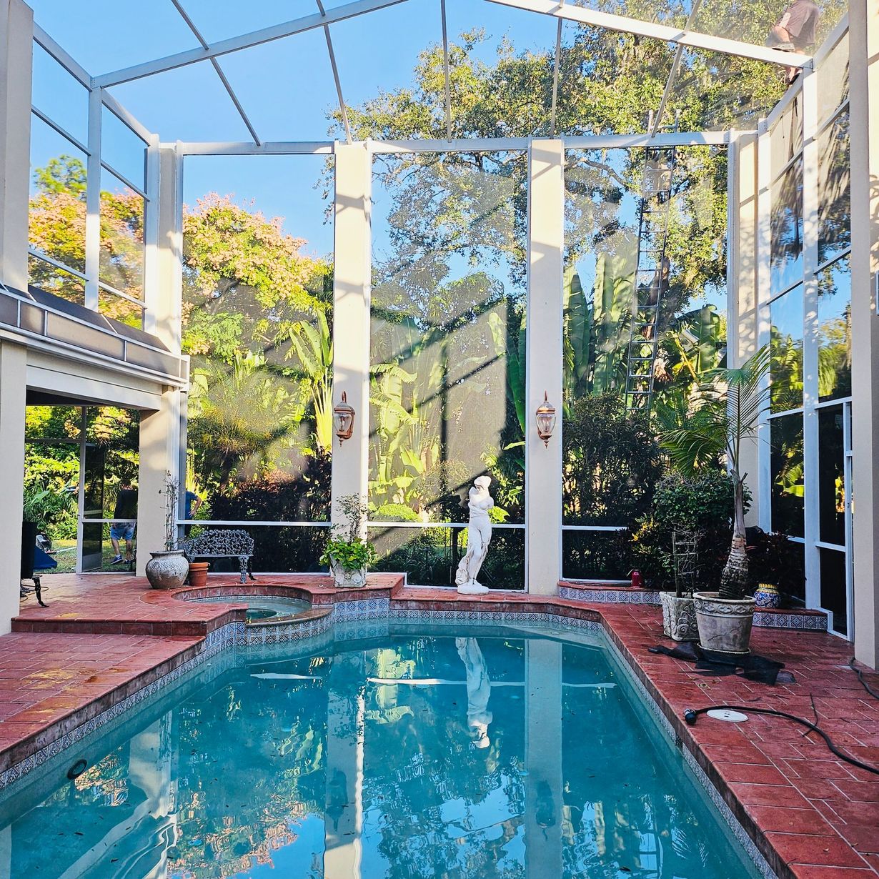 Screened backyard pool with red deck, white columns, and lush trees reflected in clear blue water
