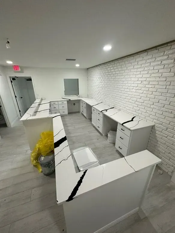 U-shaped office desks with white cabinets and marble-look countertops in a modern room with a brick accent wall.