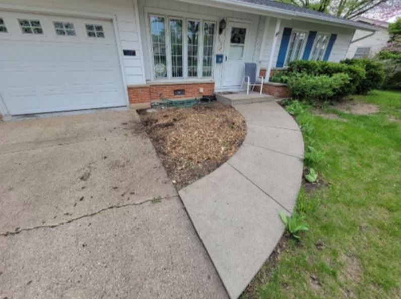 A concrete walkway leading to a house entrance with an adjacent area of mulch in front of a garage and white siding.