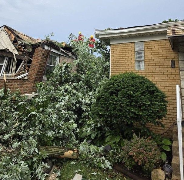 Damaged house with a collapsed roof beside a brick home and fallen tree debris after a storm