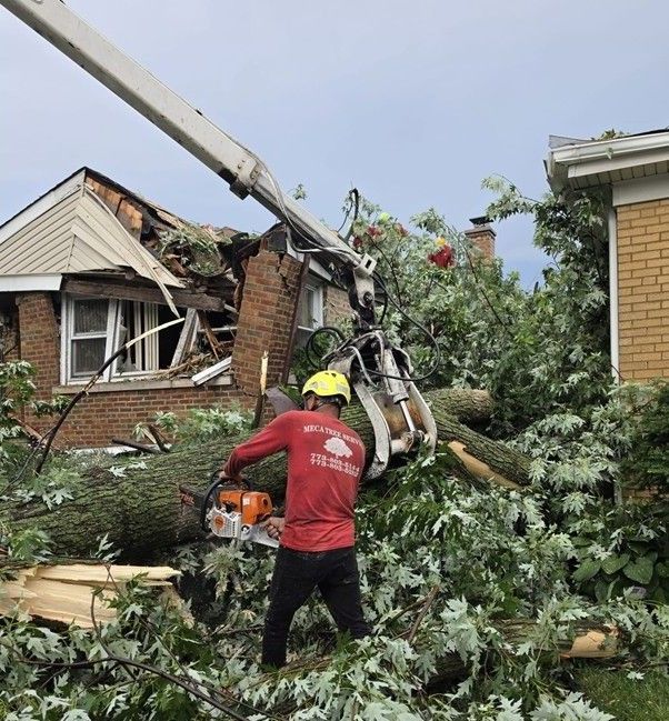 Worker in a yellow helmet clearing a fallen tree beside a damaged brick house with a crane truck