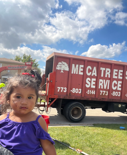 A young child in a purple shirt stands in front of a red tree service truck on a sunny day.