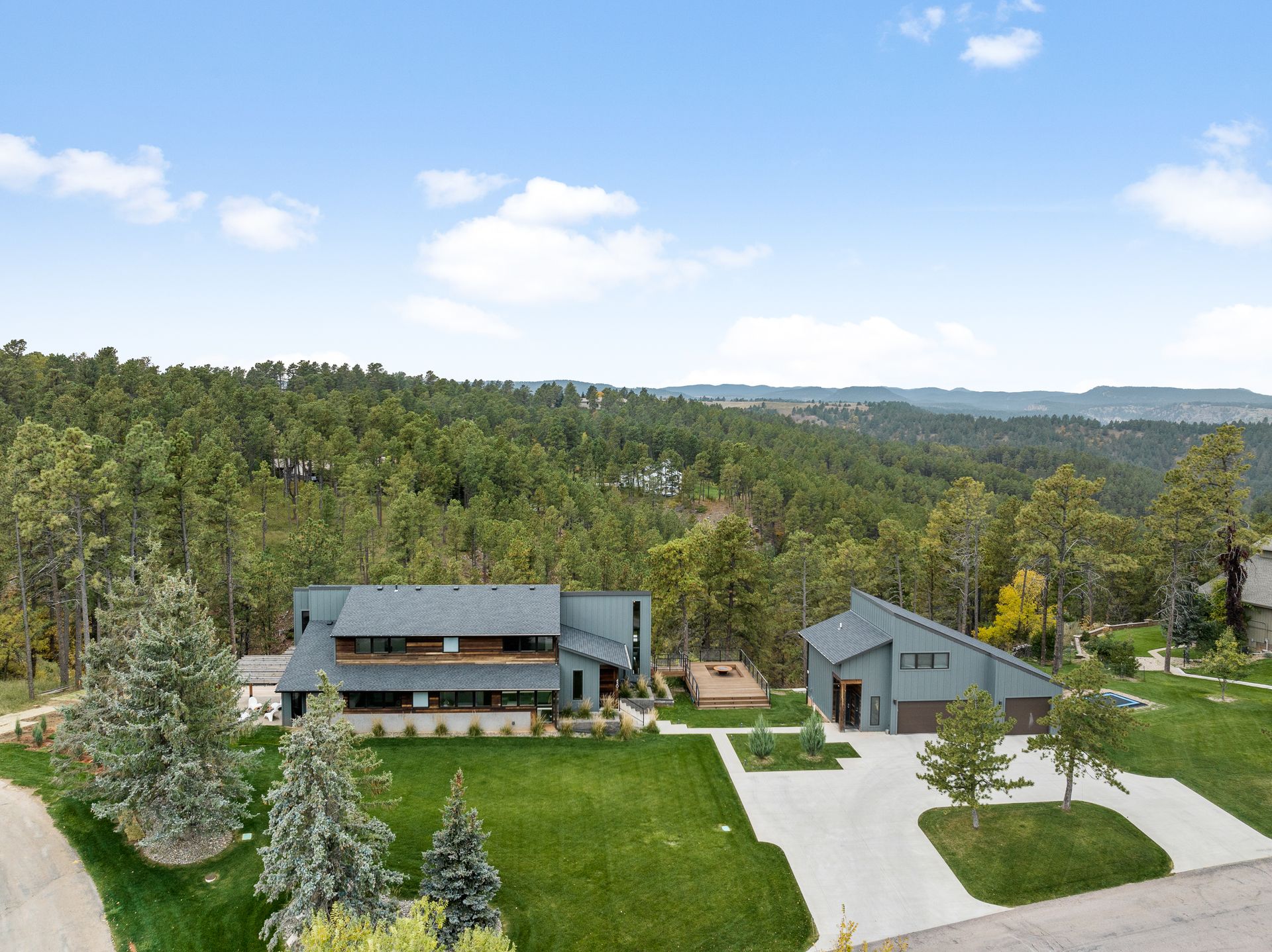 Two modern grey houses with manicured lawns against a forest backdrop under a blue sky.