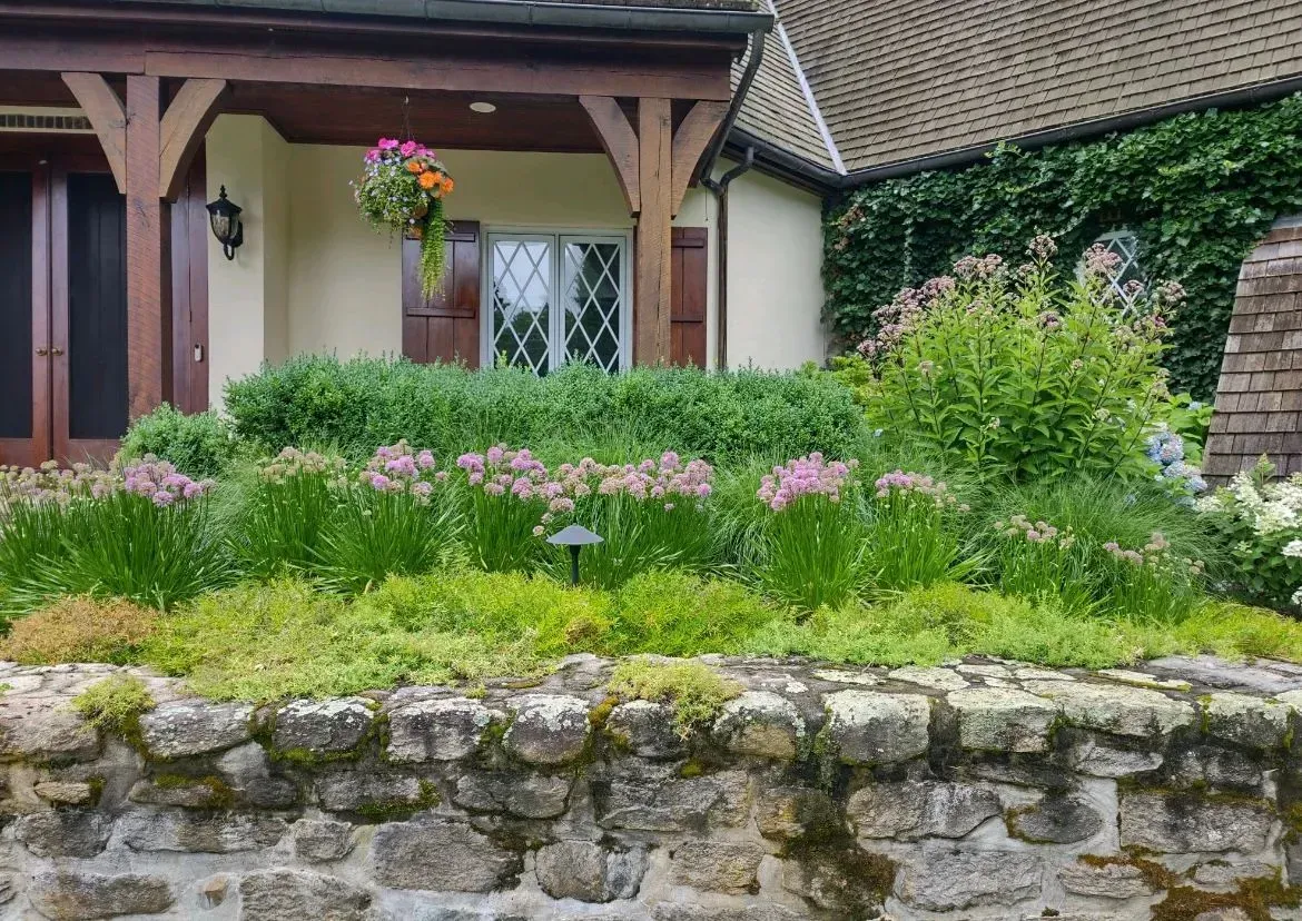 Stone wall with a garden of pink flowers and lush greenery in front of a house.