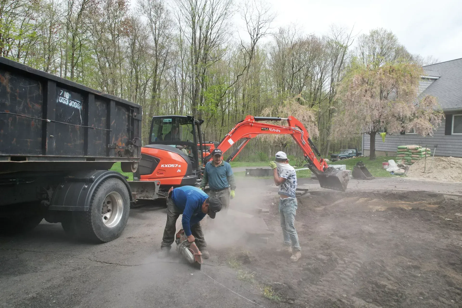 Construction workers using power tools next to an excavator and a dump trailer in an outdoor setting.
