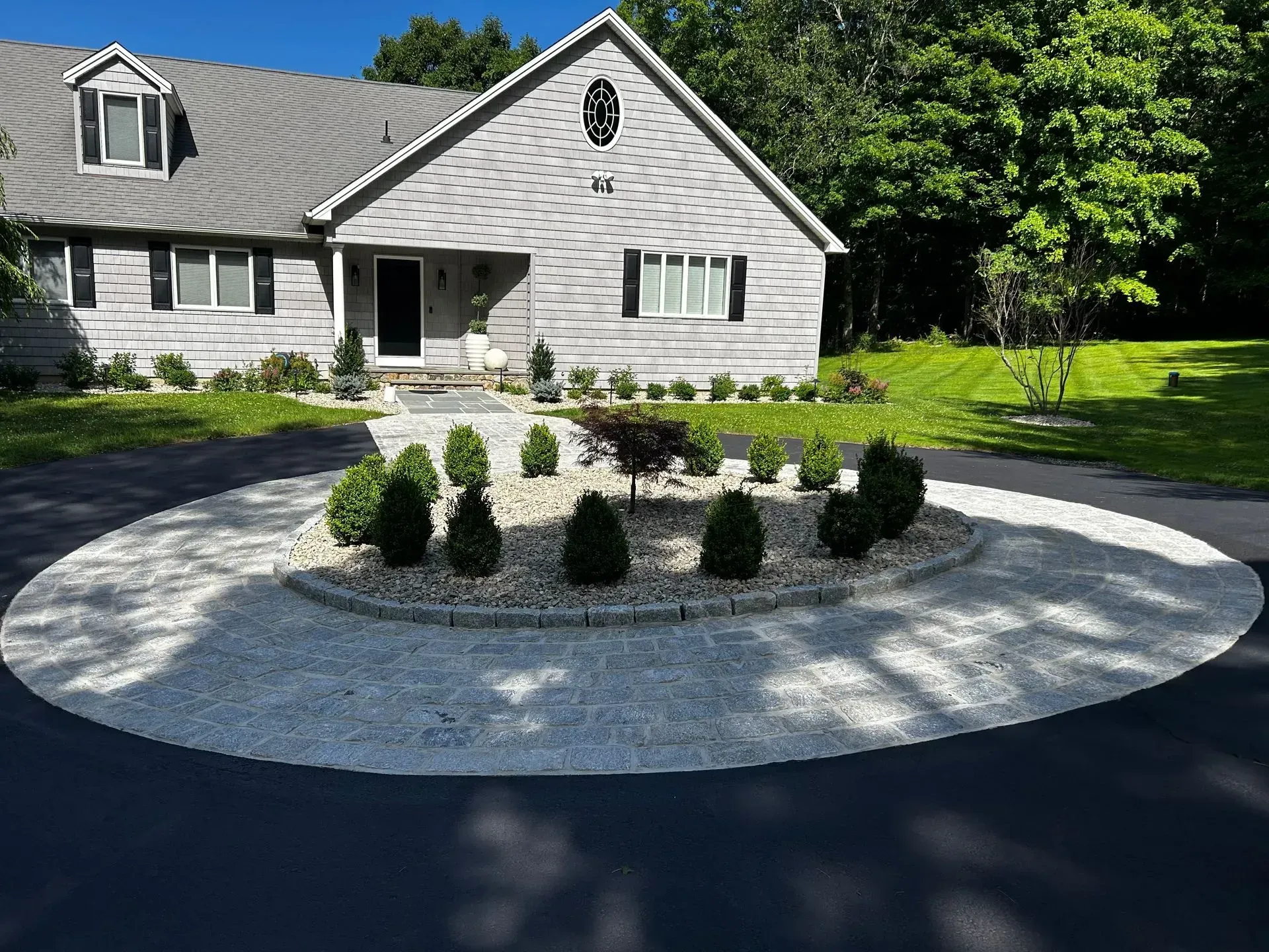 House with gray siding and circular driveway with gravel landscaping and shrubs.