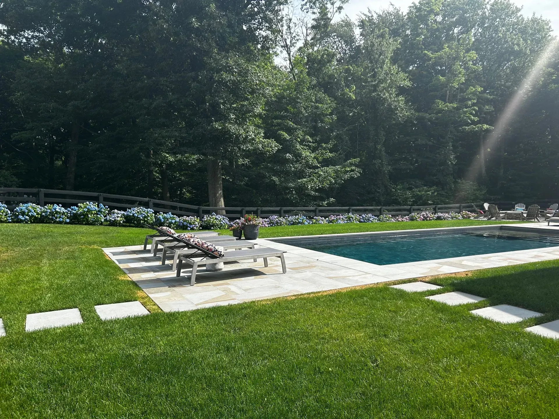 Lounge chairs by a pool on a grassy lawn, with hydrangeas and trees in the background. Sunny day.