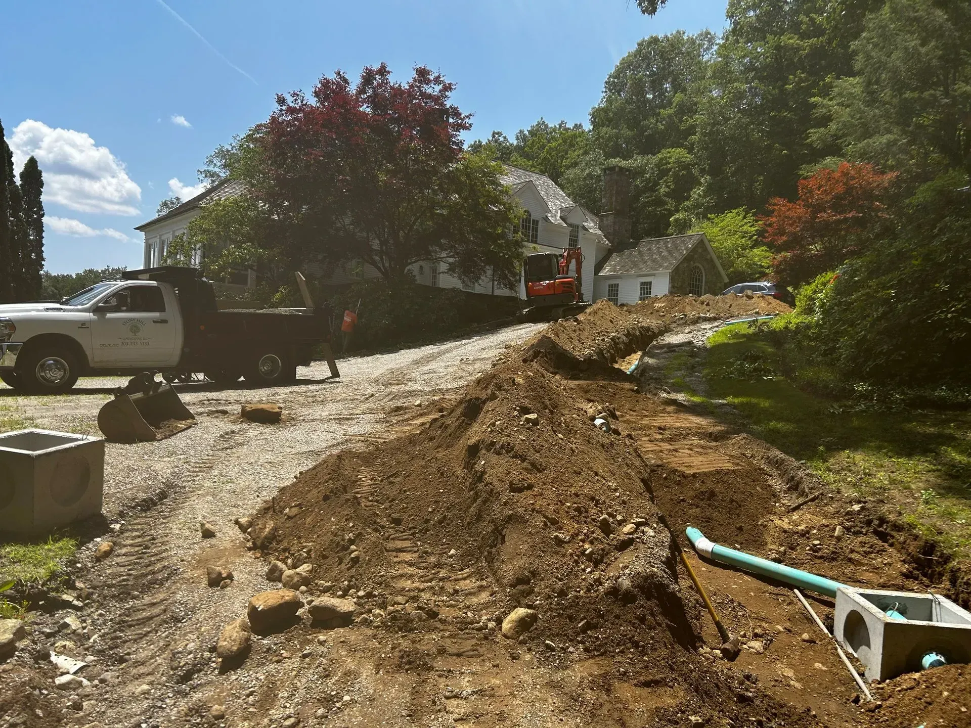 Construction site with truck, excavator, and trenching in front of a white house on a sunny day.