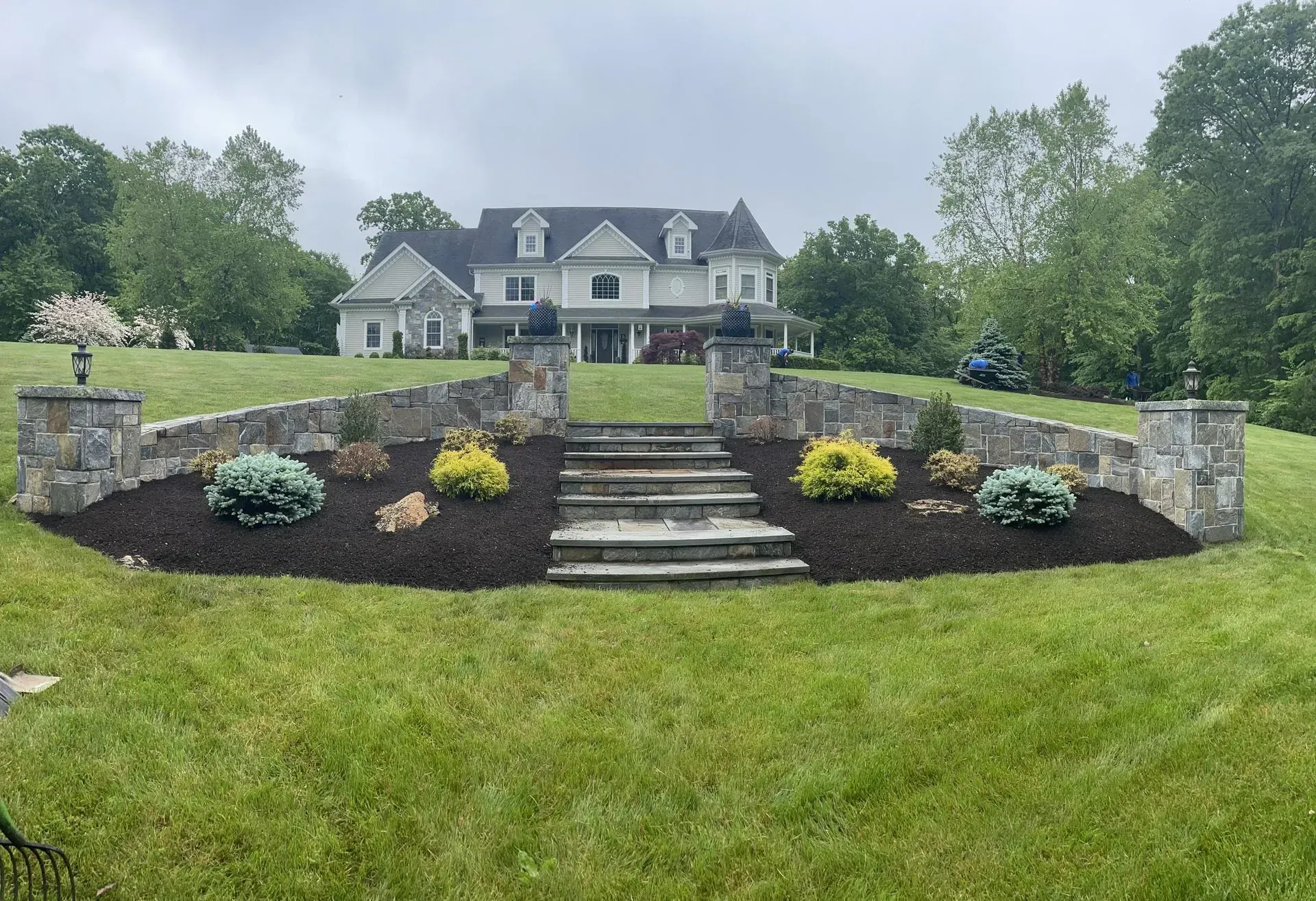 Stone wall and steps leading to a large house on a green lawn. Landscaping with bushes and mulch.
