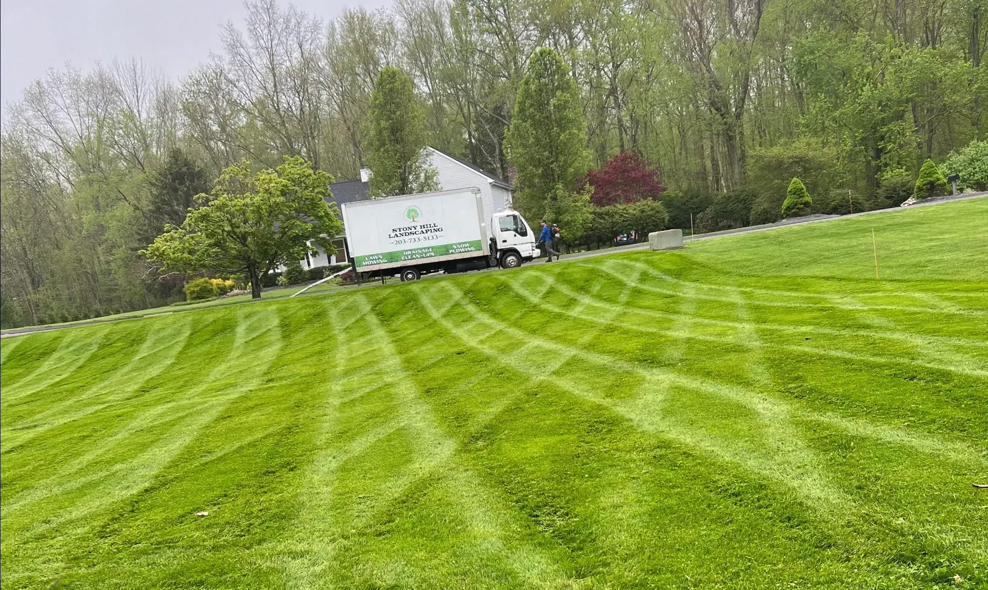 A lawn with a diamond-patterned mow, featuring a service truck parked on a grassy hill in front of trees.