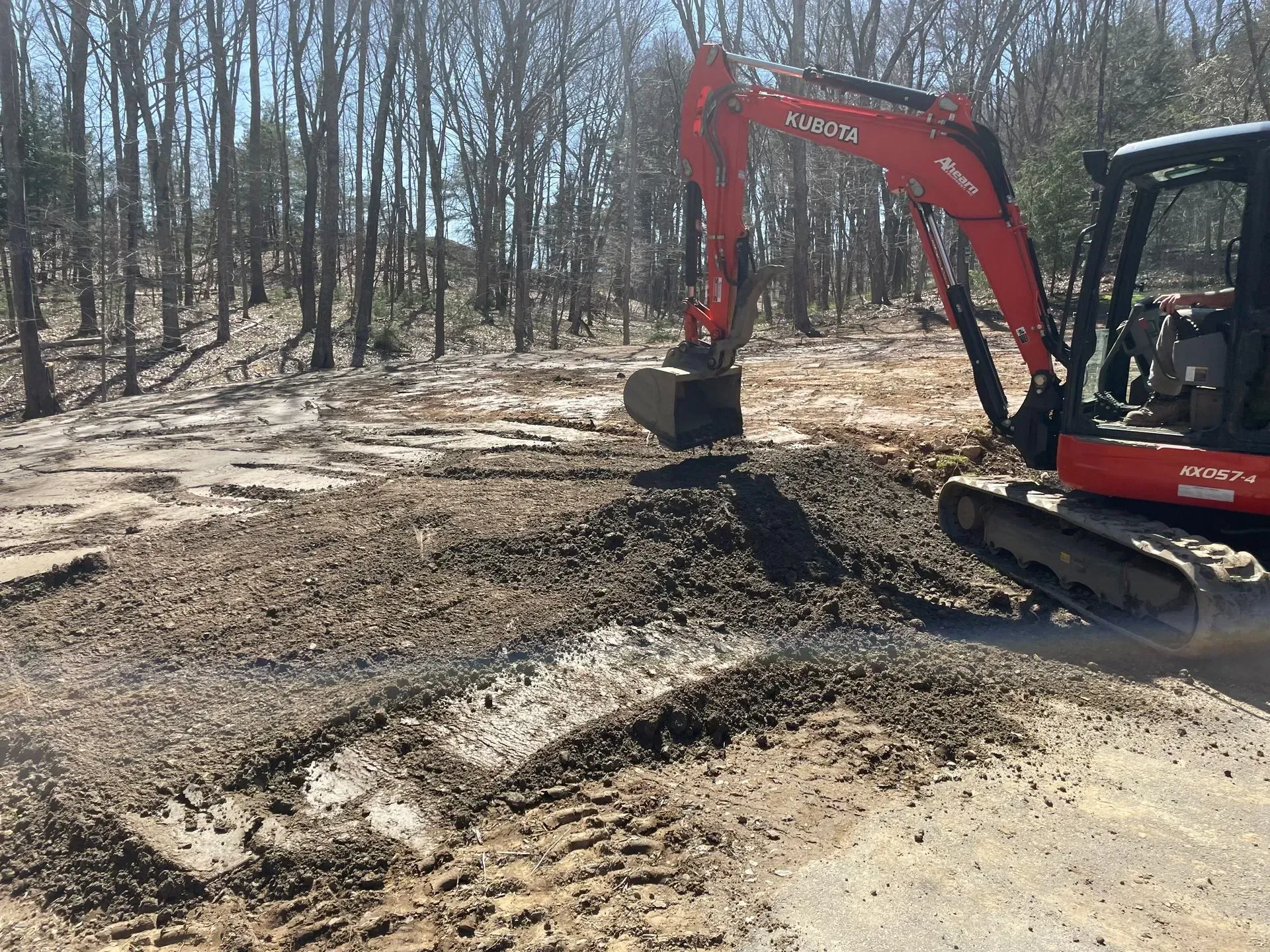 An orange Kubota excavator parked on a dirt lot in a wooded area, with an active dig site nearby.