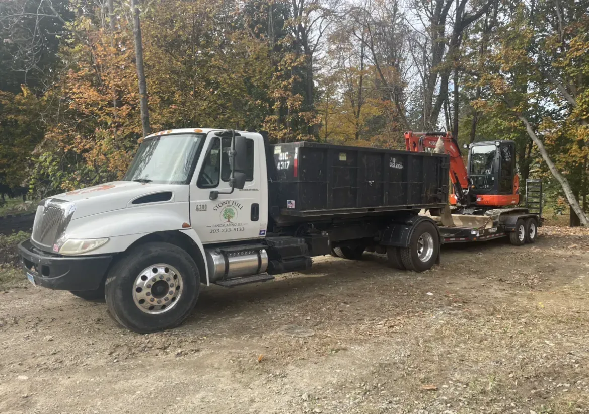 White dump truck towing an excavator on a trailer in a wooded area.