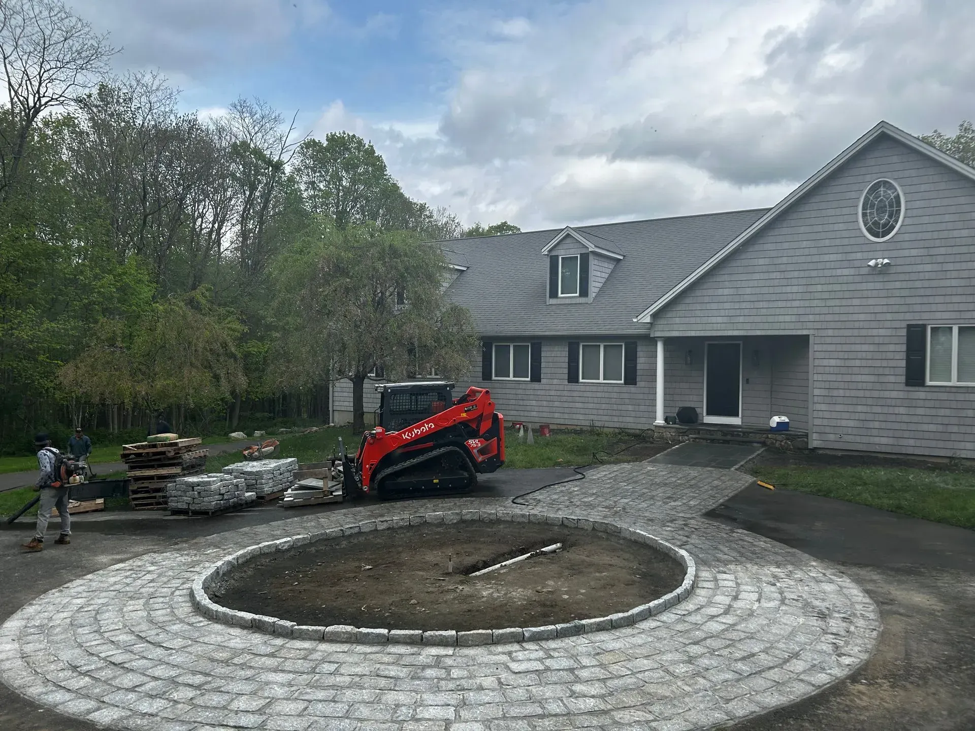 Construction site with a red skid steer, cobblestone driveway, and gray house under a cloudy sky.