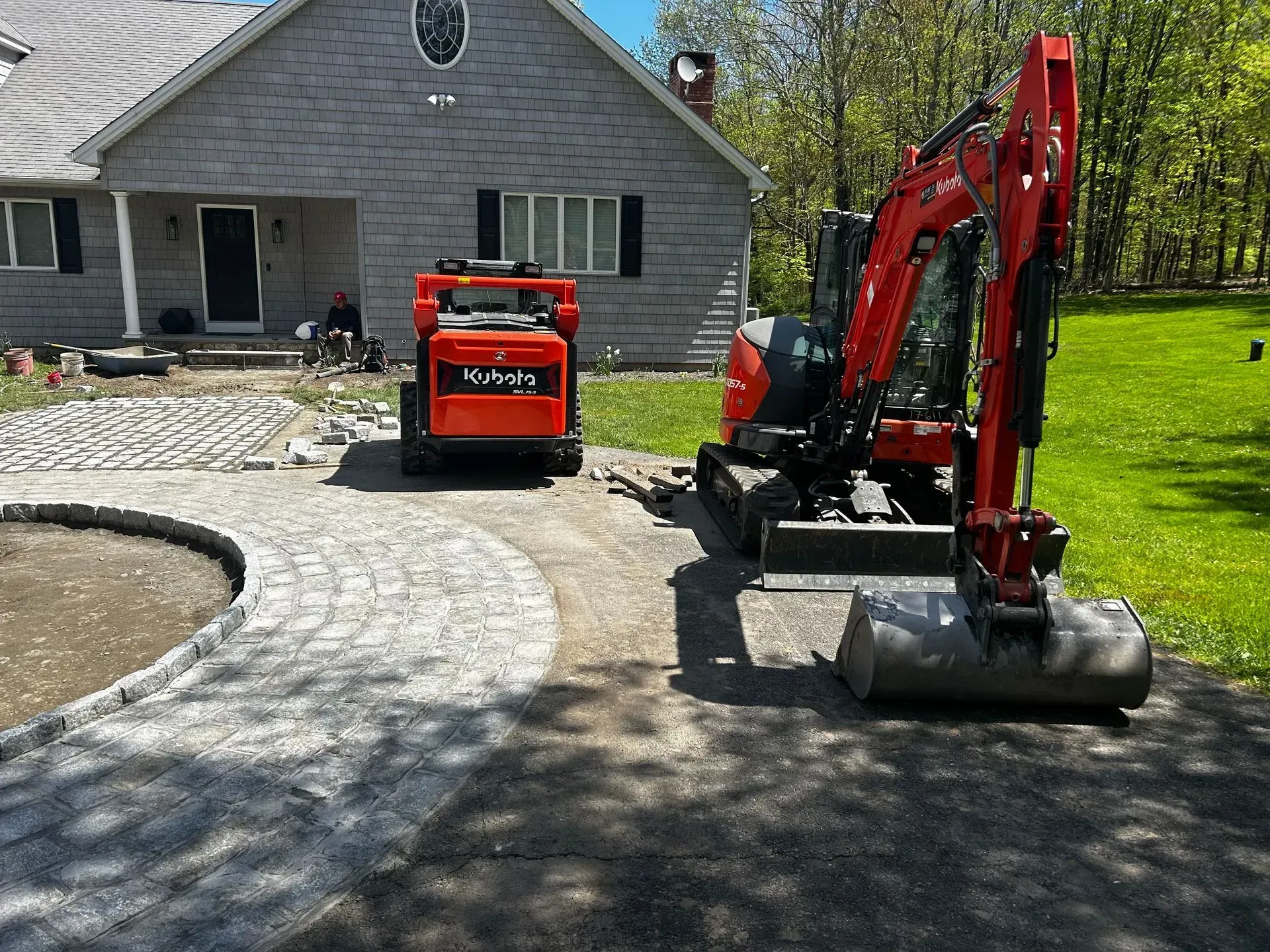 Construction site: Red excavator and dumper working on a brick paved driveway in front of a house.