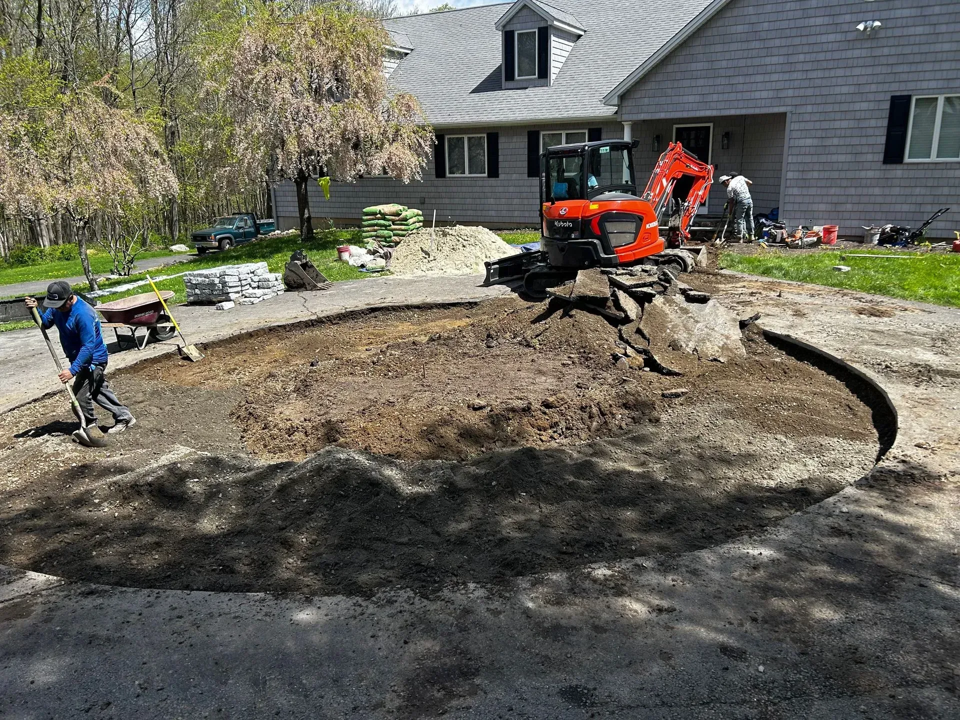 A worker uses a shovel near an orange excavator digging up a circular dirt area in front of a house.