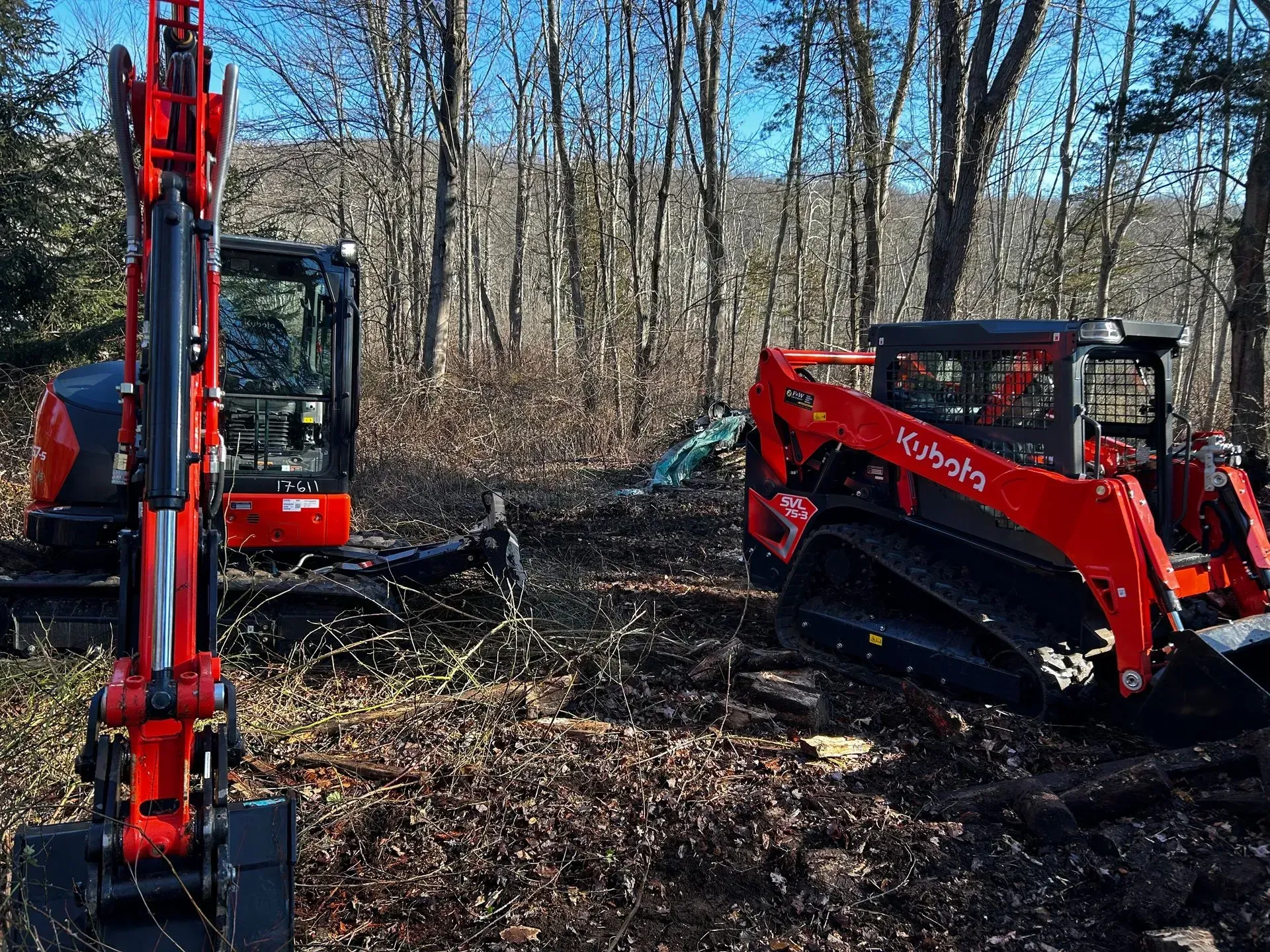 A red Kubota compact excavator and a red Kubota track loader sit in a wooded area surrounded by cleared brush.