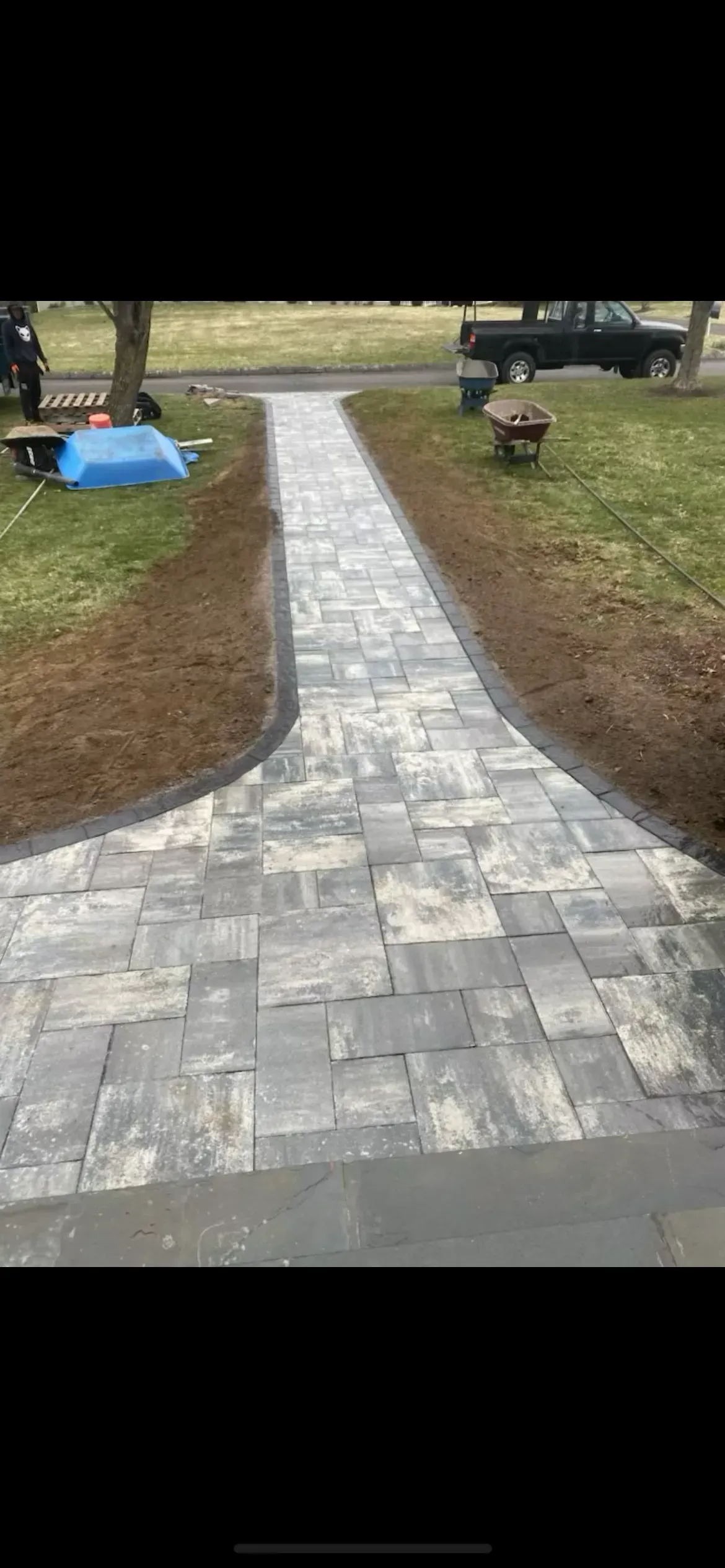 A newly installed gray paver walkway leads from a green lawn toward a street with a parked truck.