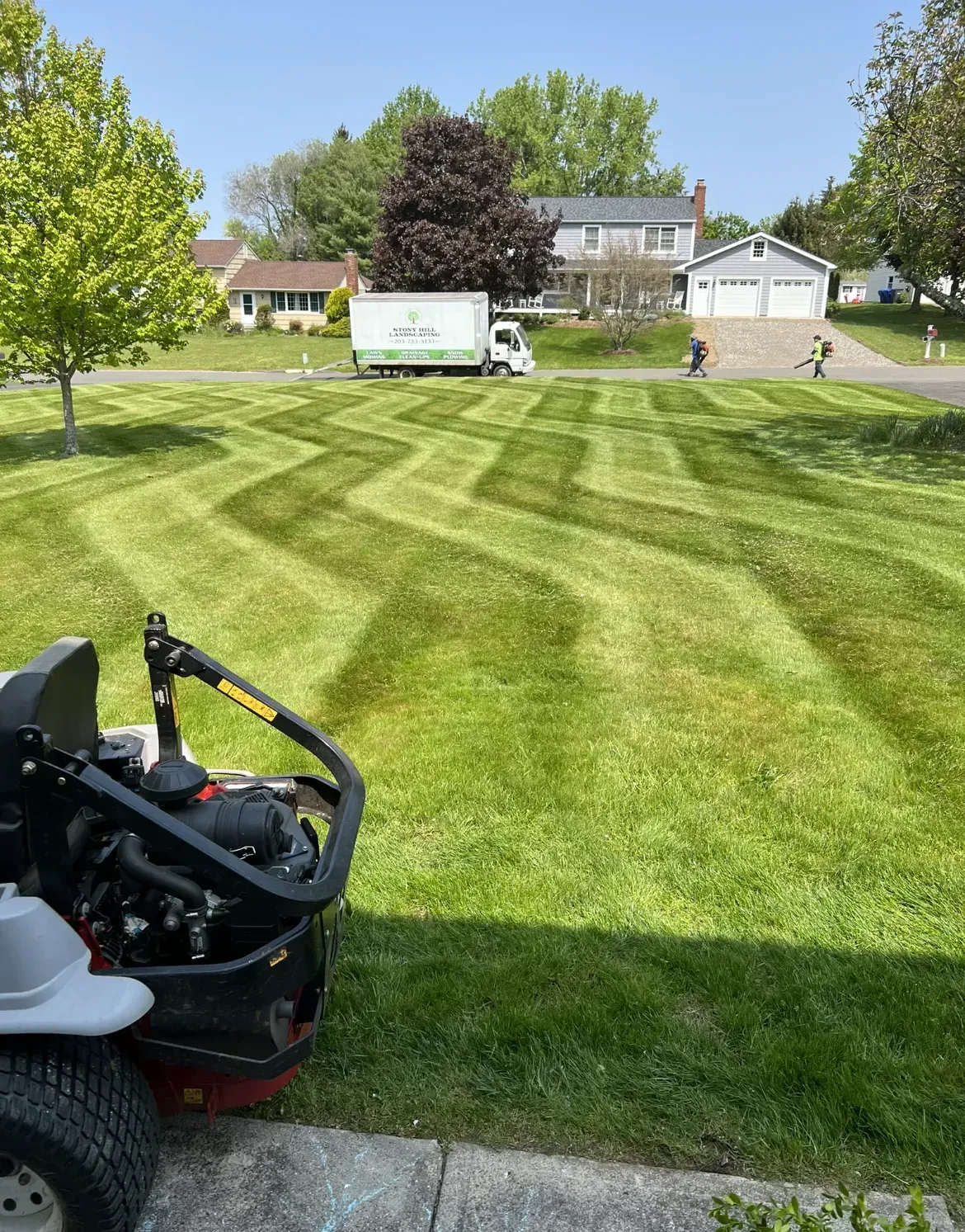 A zero-turn lawn mower overlooks a grassy yard with zig-zag mow patterns, with a service truck and houses in the distance.