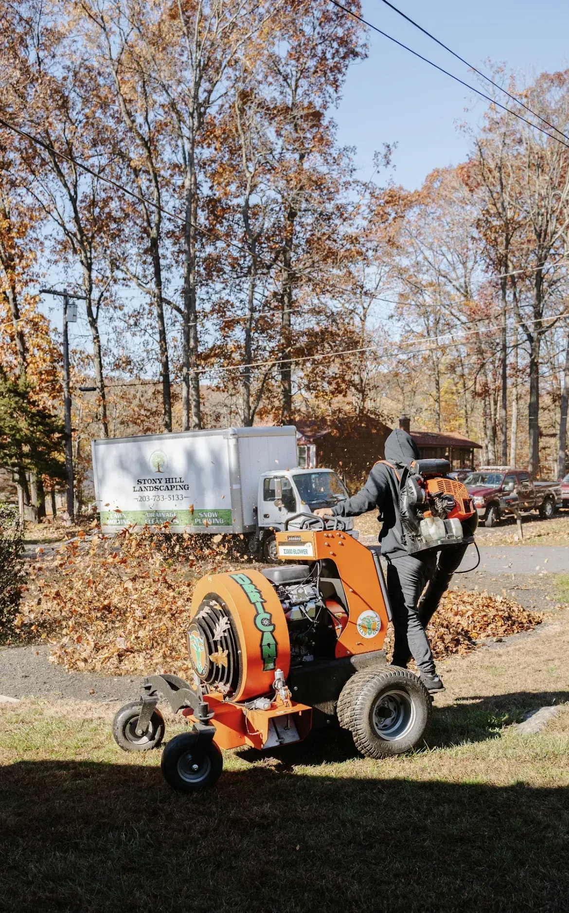 A person in dark clothing uses a large orange leaf blower on a grassy lawn with autumn trees and a trailer in the back.