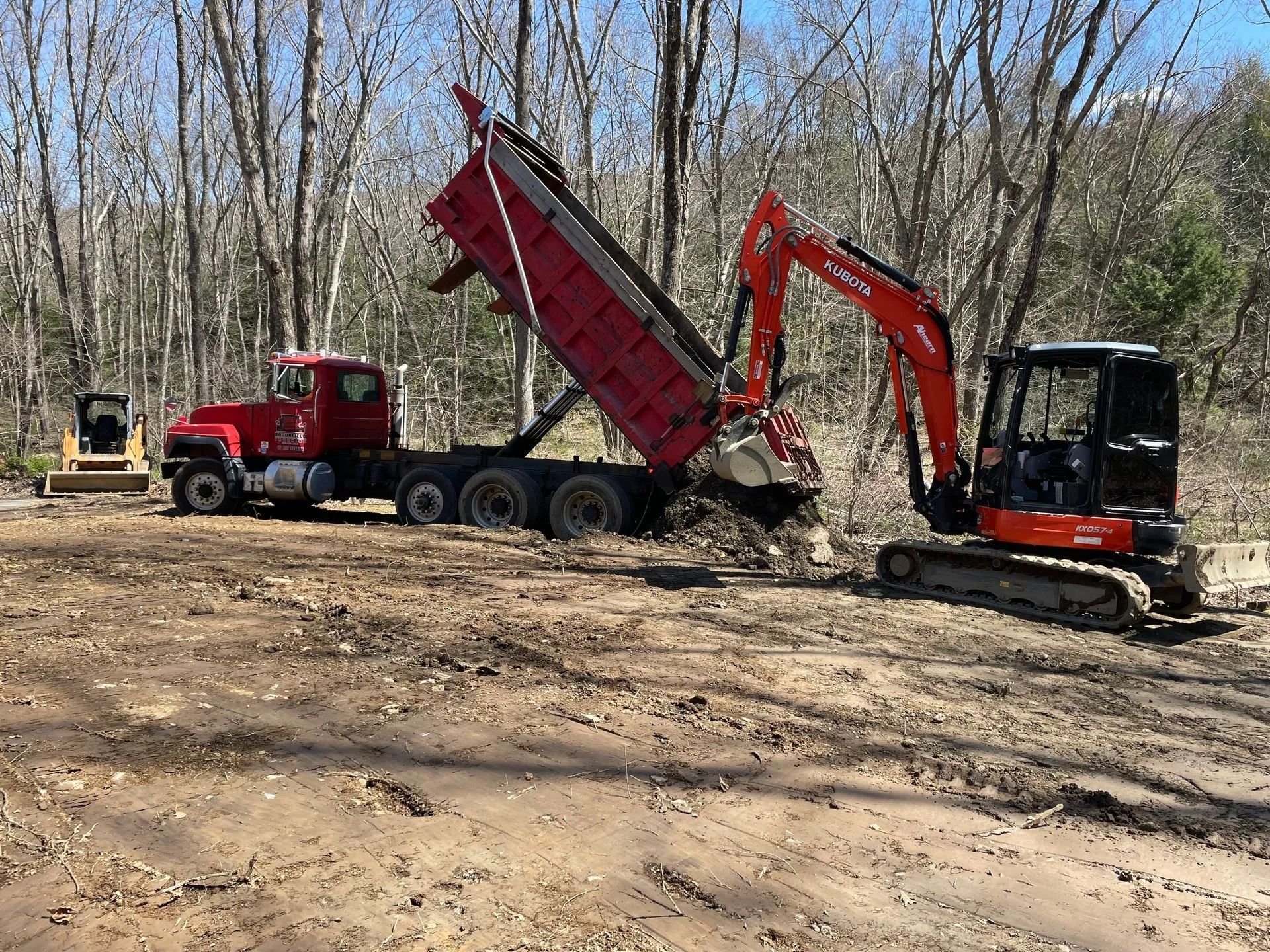 An orange excavator operates near a red dump truck on a dirt construction site surrounded by leafless trees.